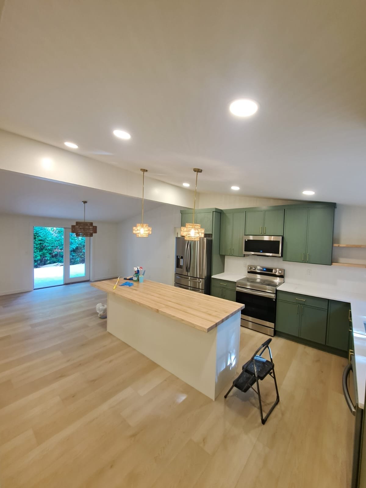 Modern kitchen with green cabinets, stainless steel appliances, a wooden island, hanging pendant lights, hardwood flooring, and a large sliding glass door leading outside.