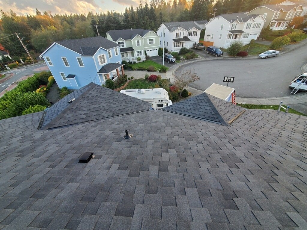 An aerial view of a neighborhood showing rooftops, yards, parked cars, and a curved street with trees and houses, with a mountain backdrop and a partly cloudy sky.