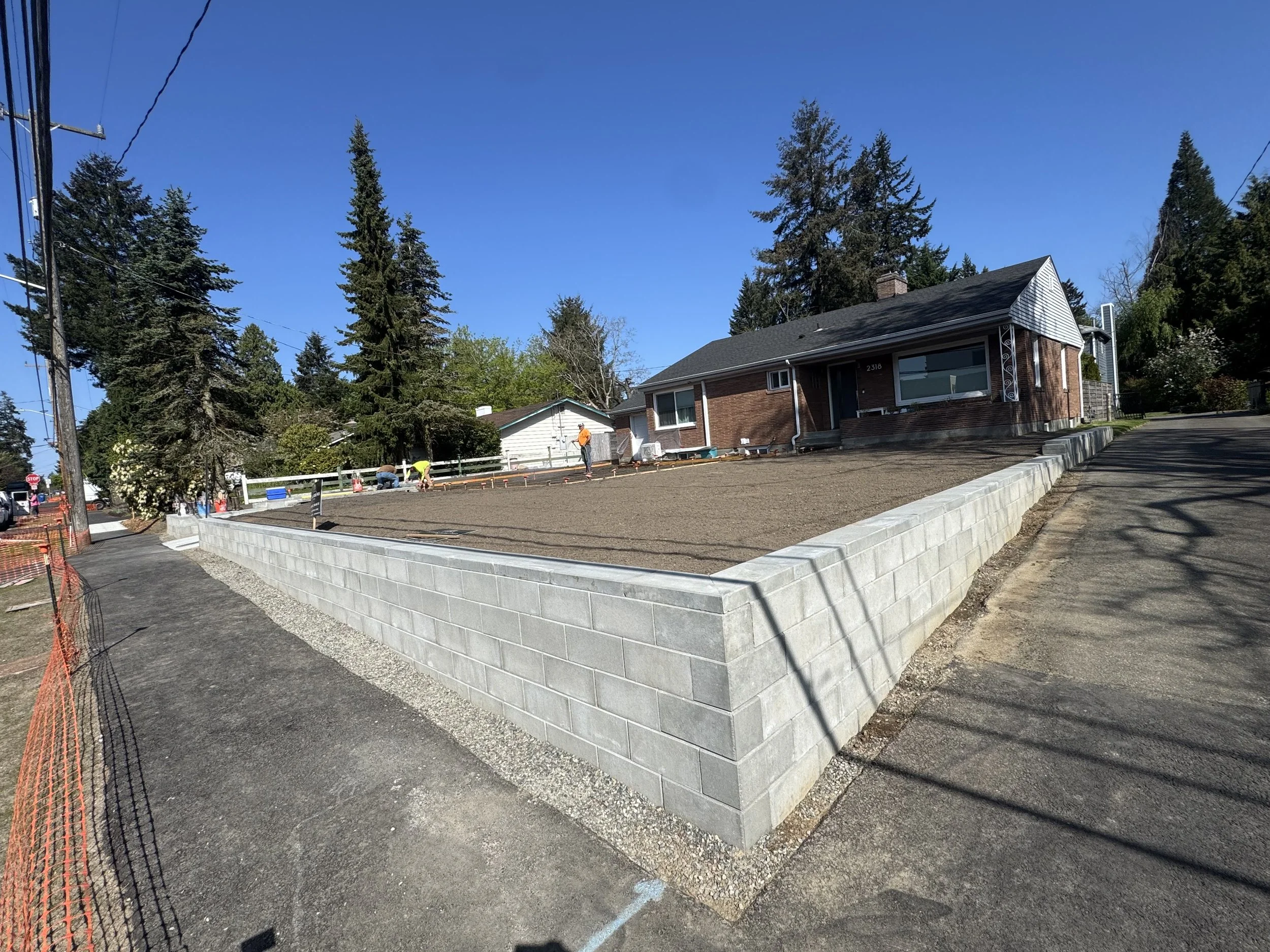 A residential construction site with a brick house in the background and a new concrete block wall being built in the foreground, with workers present and a blue sky overhead.
