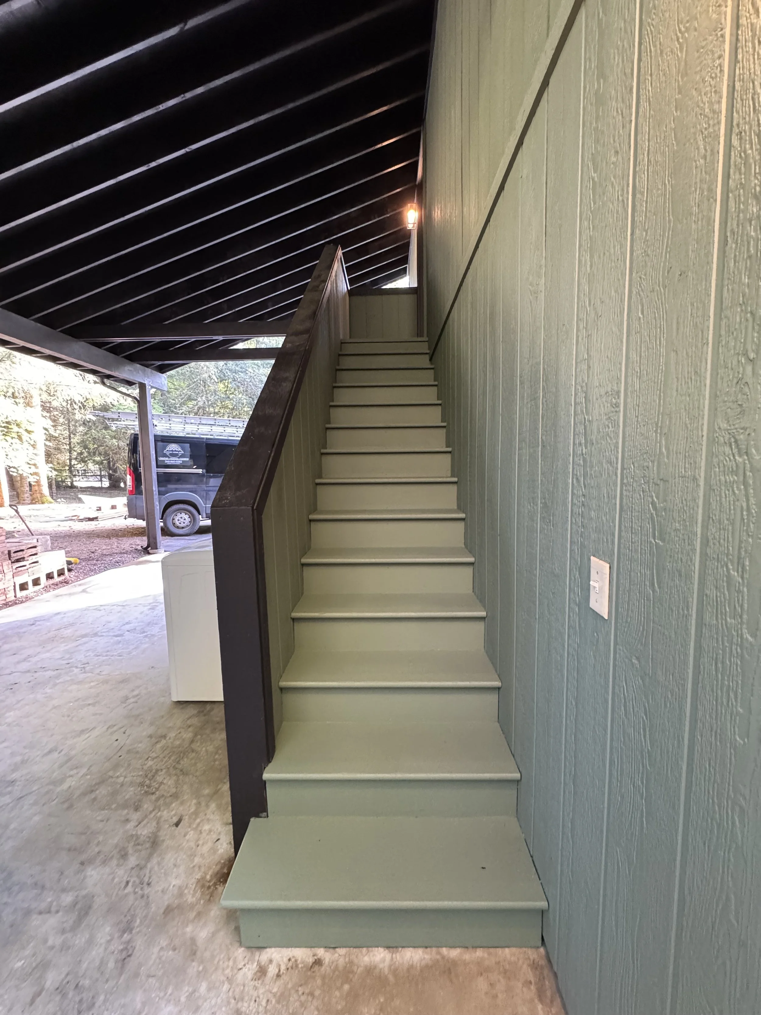Green staircase with dark railing against a green textured wall, with an outdoor view of trees and a black van in the background.