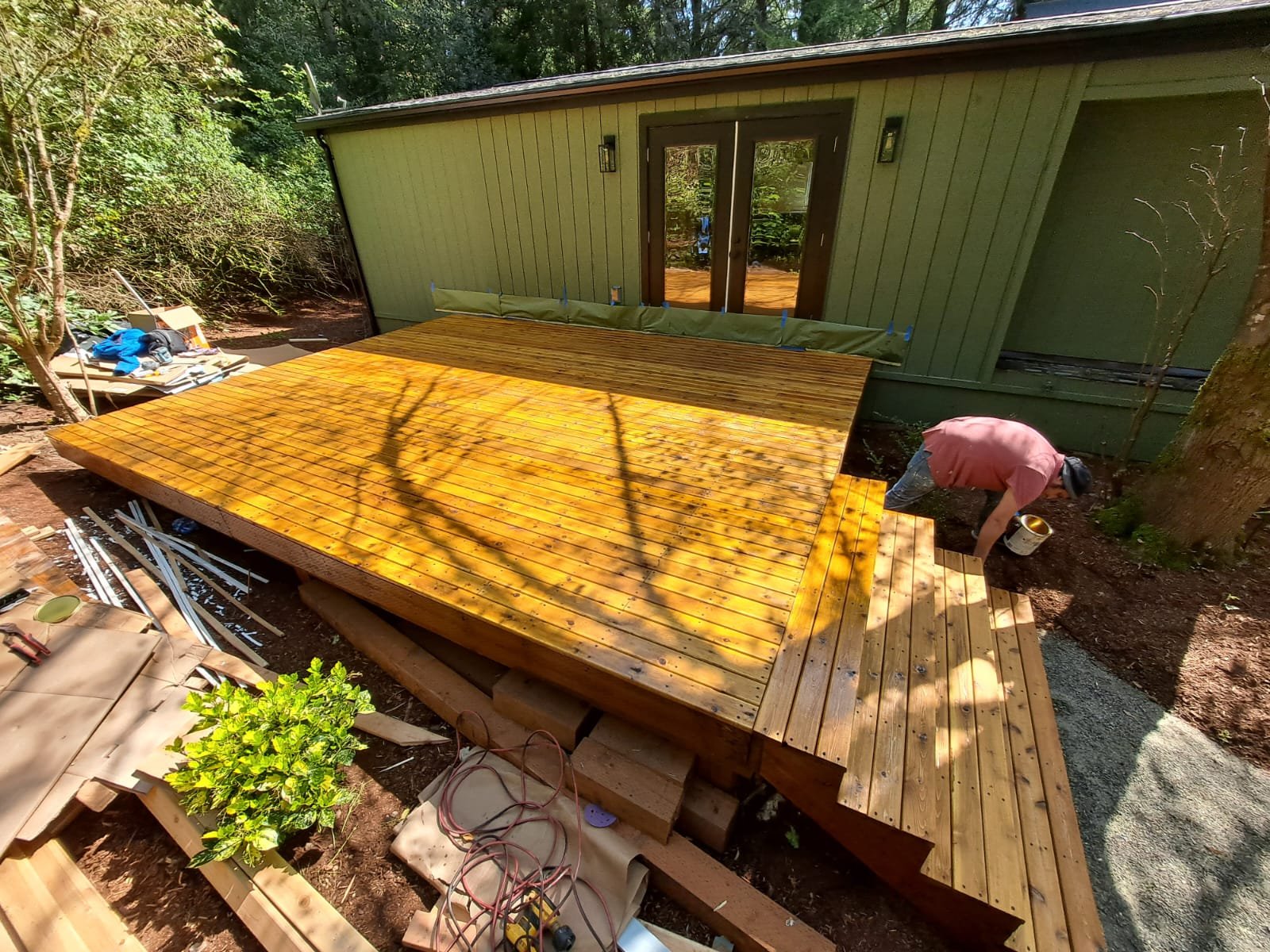 Construction worker installing a wooden deck on the backyard of a green house surrounded by trees.