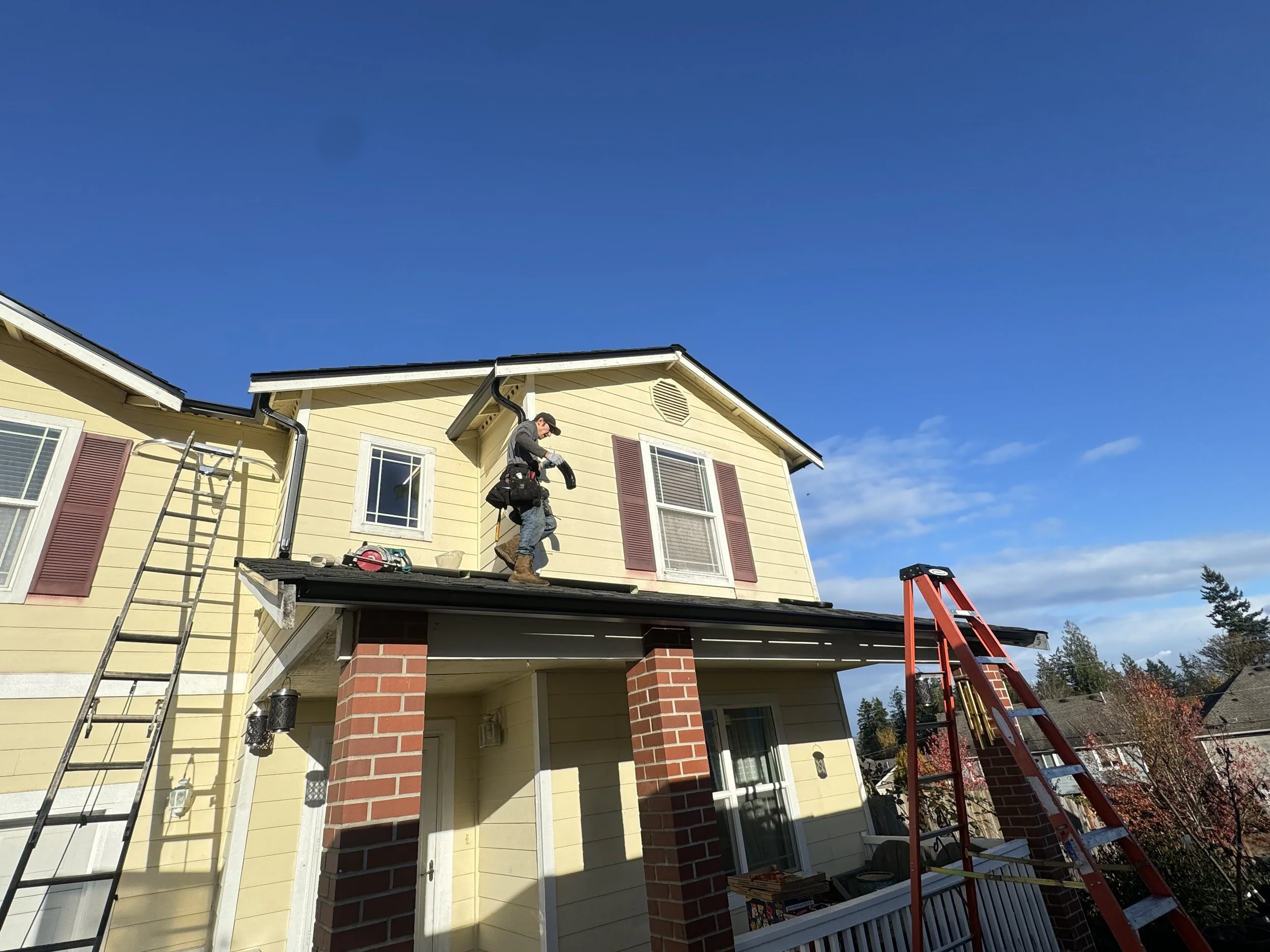 Construction worker standing on the roof of a yellow house with red shutters, installing or repairing the roof under a clear blue sky. A red ladder leans against the house and a black ladder is positioned on the side.
