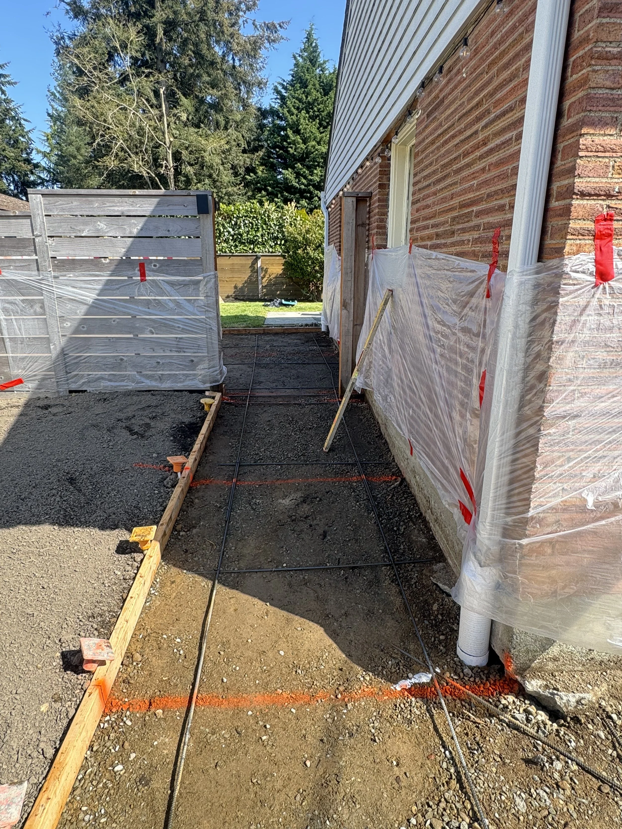 Construction site with a partially paved walkway beside a brick house, plastic sheeting covering windows, and construction markings on the ground, with trees and a clear blue sky in the background.