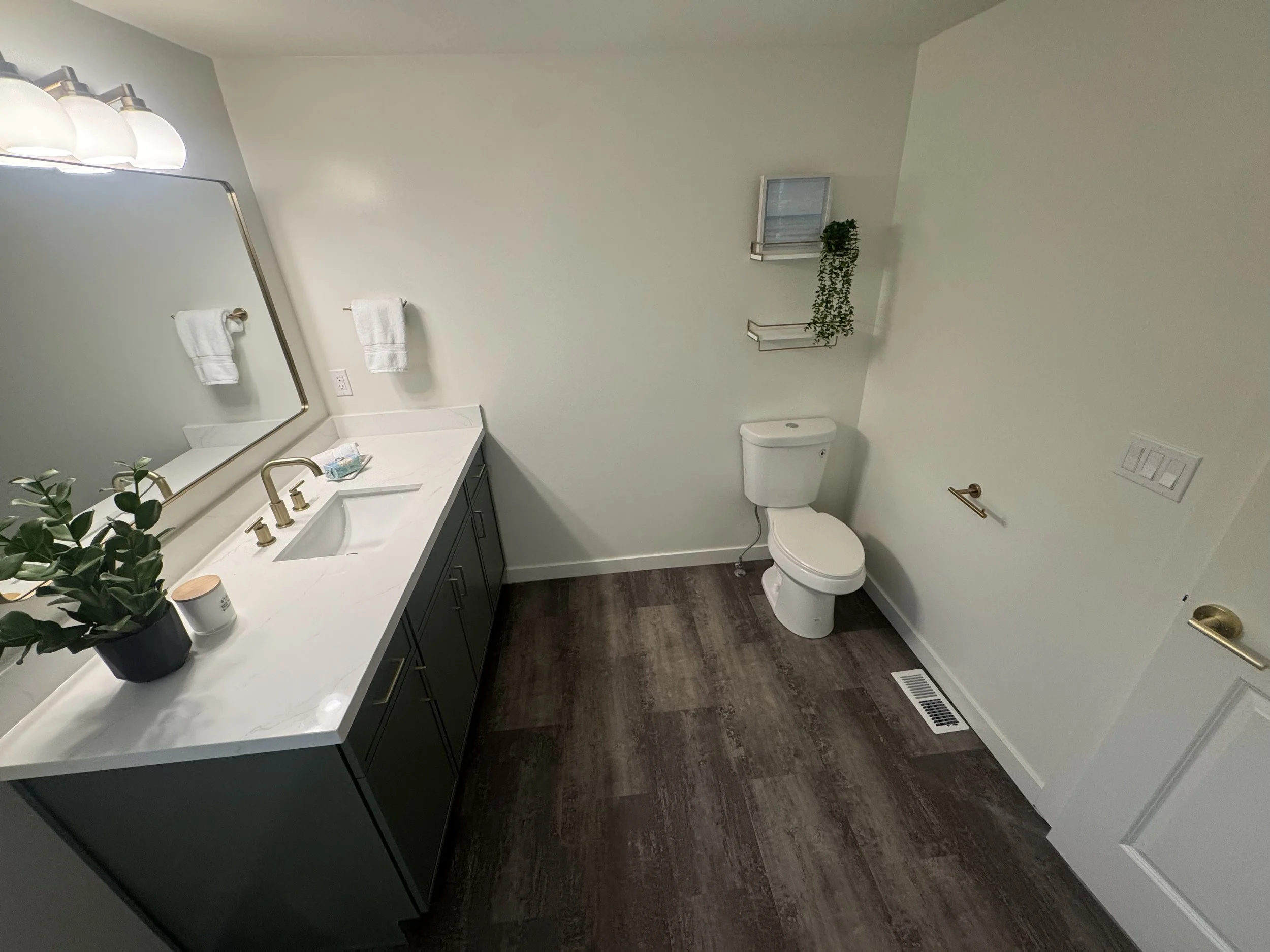 Modern bathroom with a white vanity, gold fixtures, a large mirror, two towel racks with white towels, a potted plant, wall shelves with greenery, a toilet, and wood-look flooring.