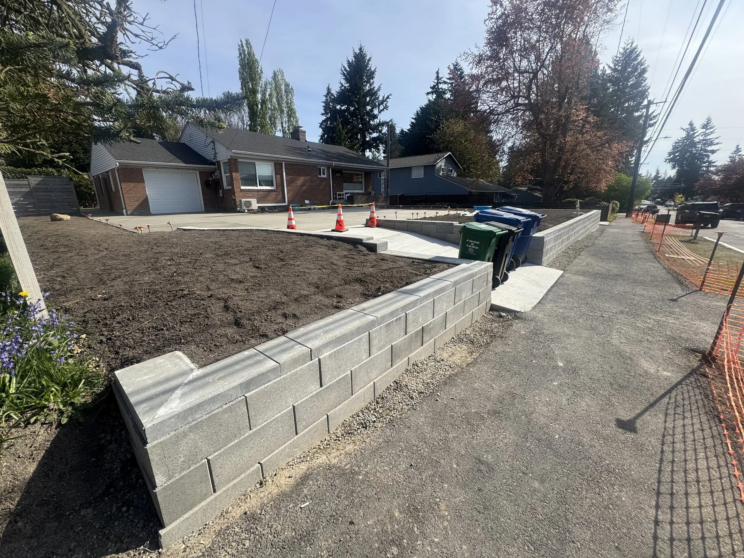Sidewalk under construction with a cinder block wall and several trash cans along the curb, with houses and trees in the background.