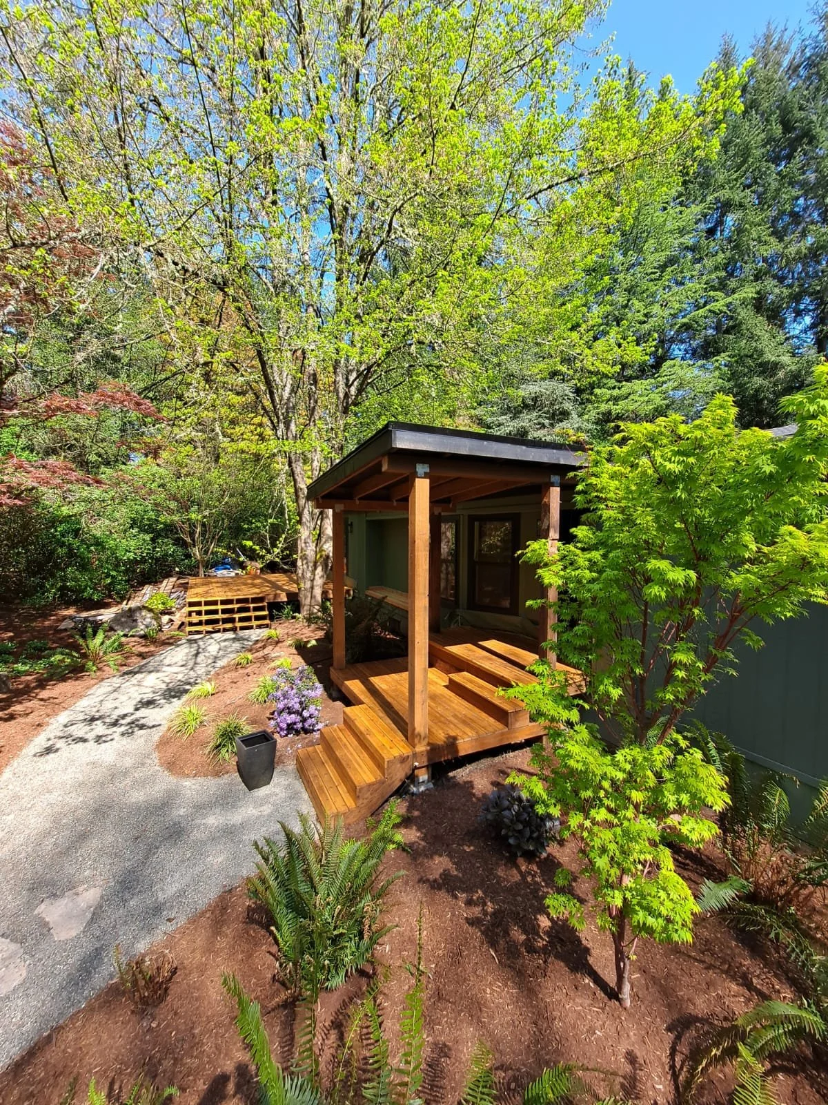 A small wooden porch with steps leads to a house with large windows, surrounded by lush green trees and plants in a garden.