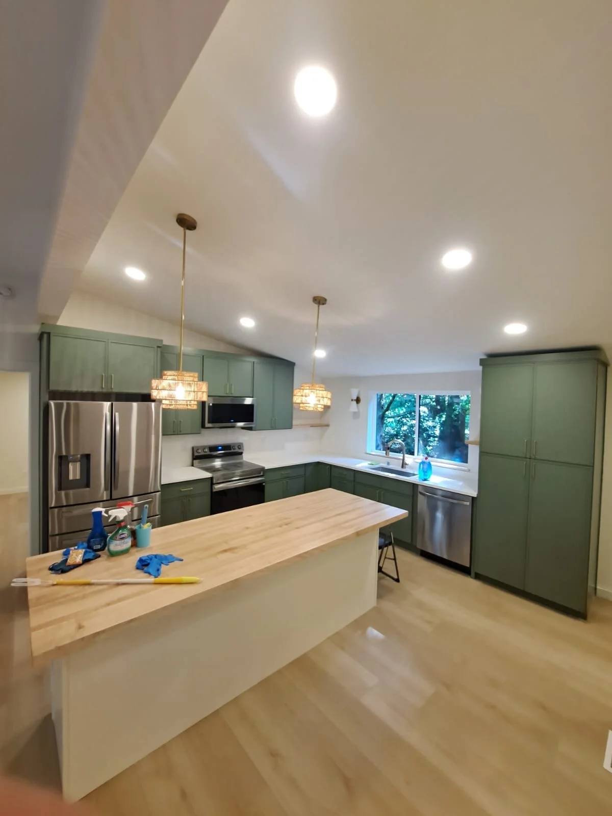 Modern kitchen with green cabinets, stainless steel appliances, a wooden island, pendant and recessed lighting, and a large window with outdoor greenery.