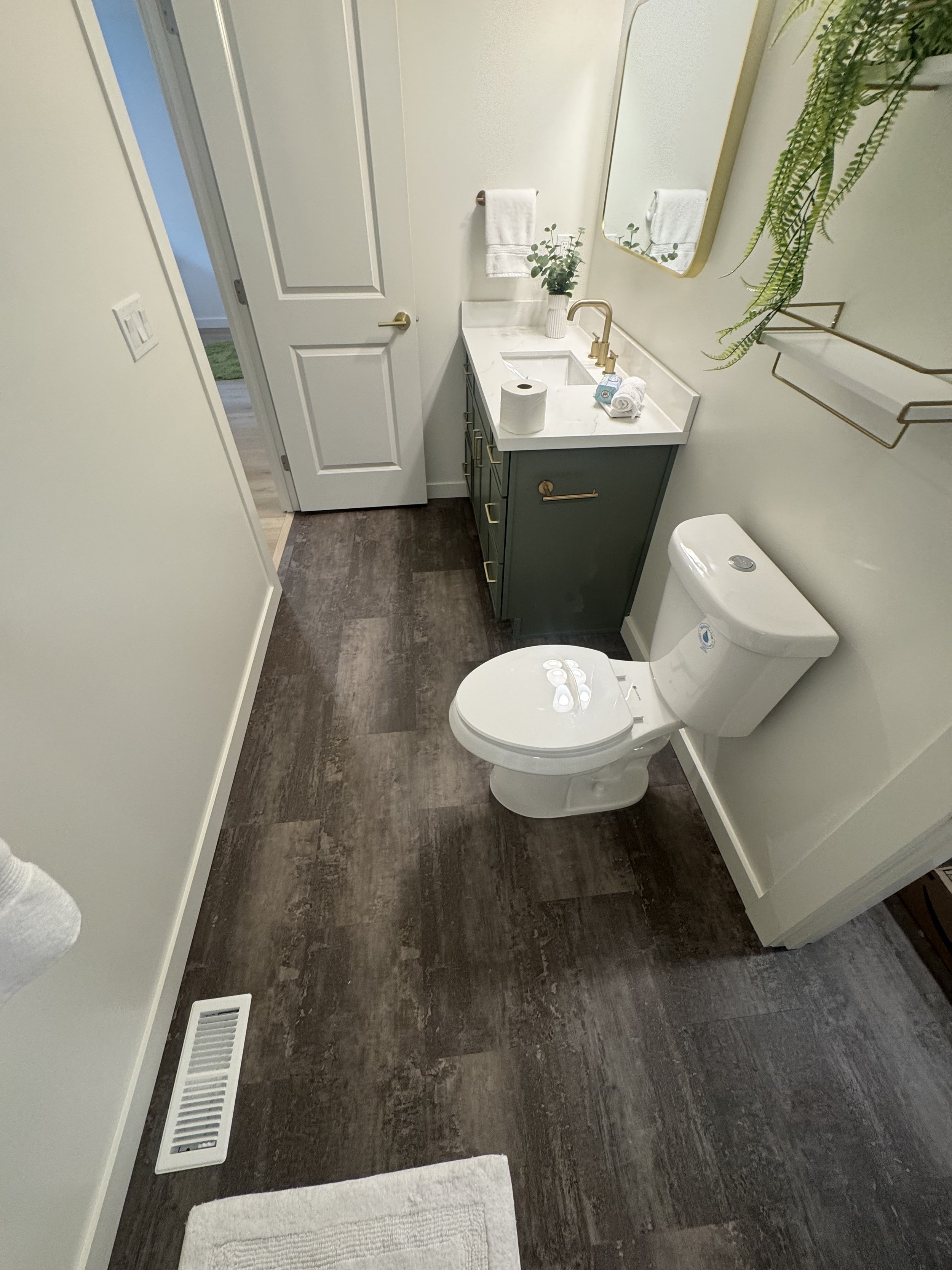 A small modern bathroom with a toilet, a green vanity with a white marble countertop, a gold faucet, and a mirror. There are white towels, a potted plant, and a gold towel rack on the wall. The bathroom has wood-look flooring and a vent on the floor.