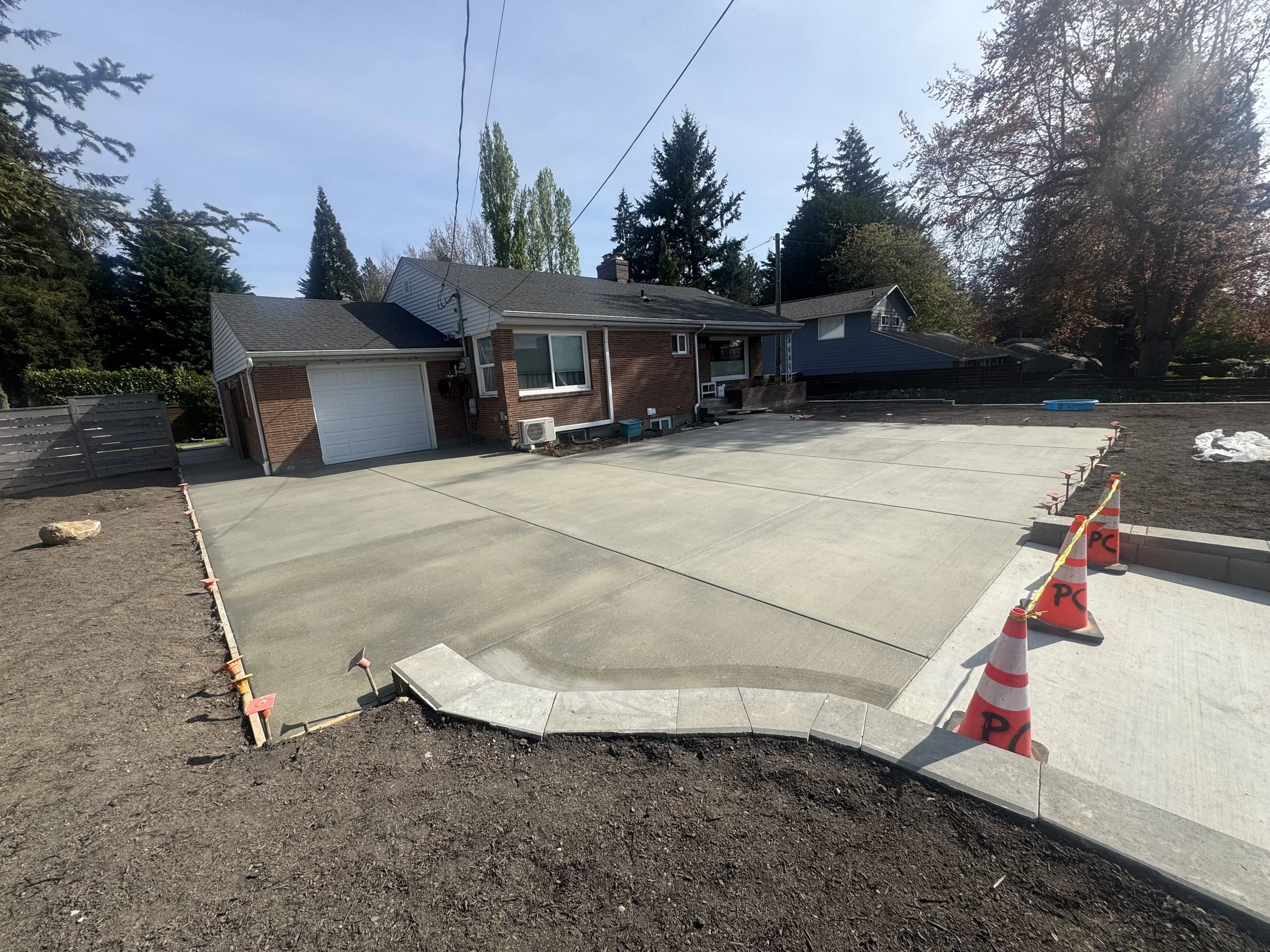 A newly poured concrete driveway in front of a brick house with a garage, with orange construction cones placed along the edge.