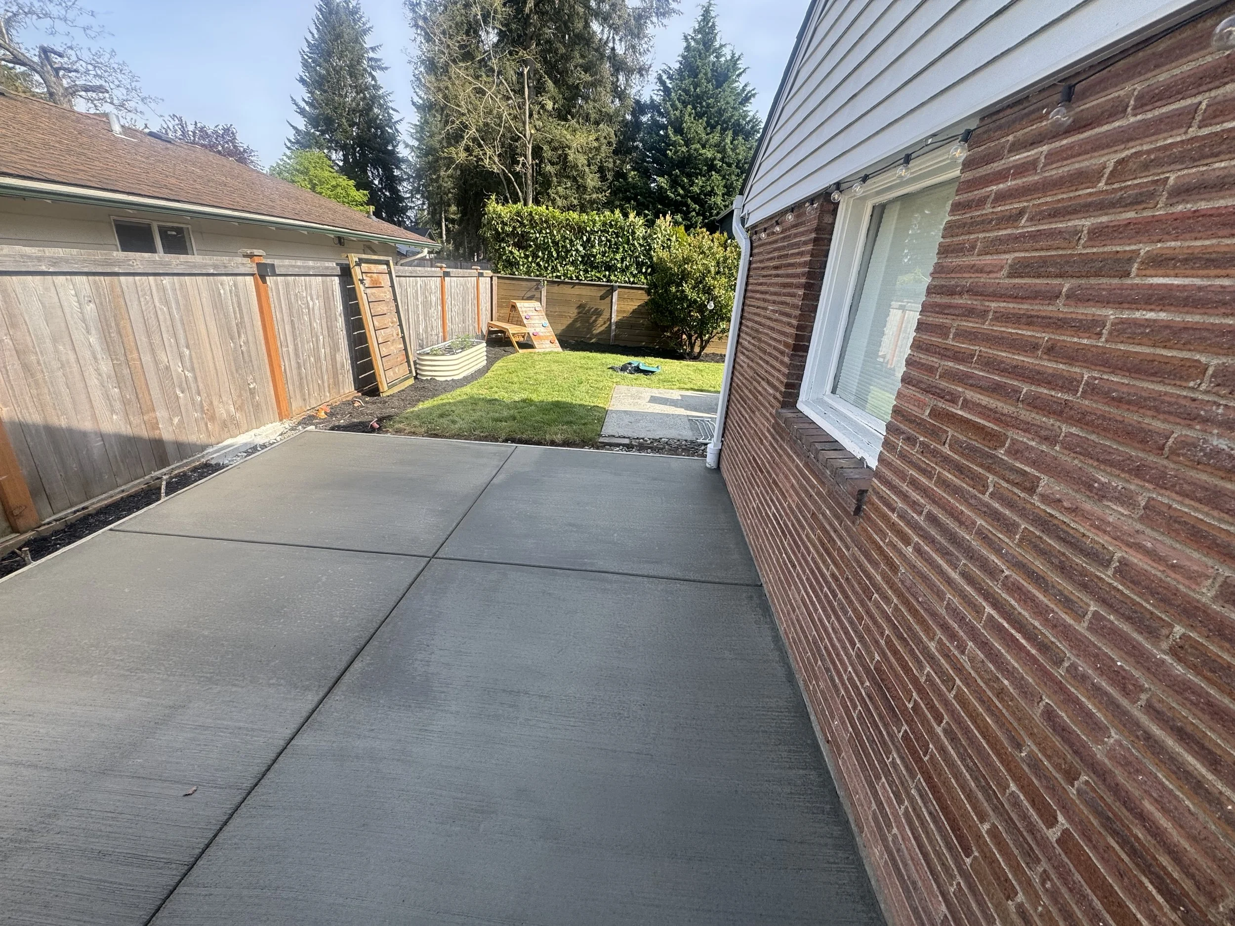 A backyard patio with concrete flooring next to a brick house, a grass yard, a wooden fence, and yard furniture with trees in the background.