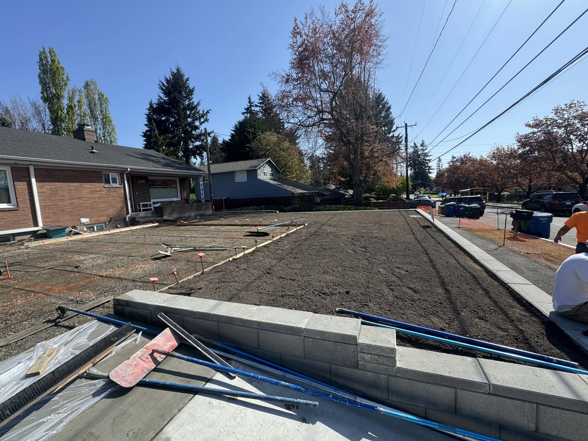 Construction site for a new sidewalk in front of a house, with equipment and materials on the ground, trees, and houses in the background.