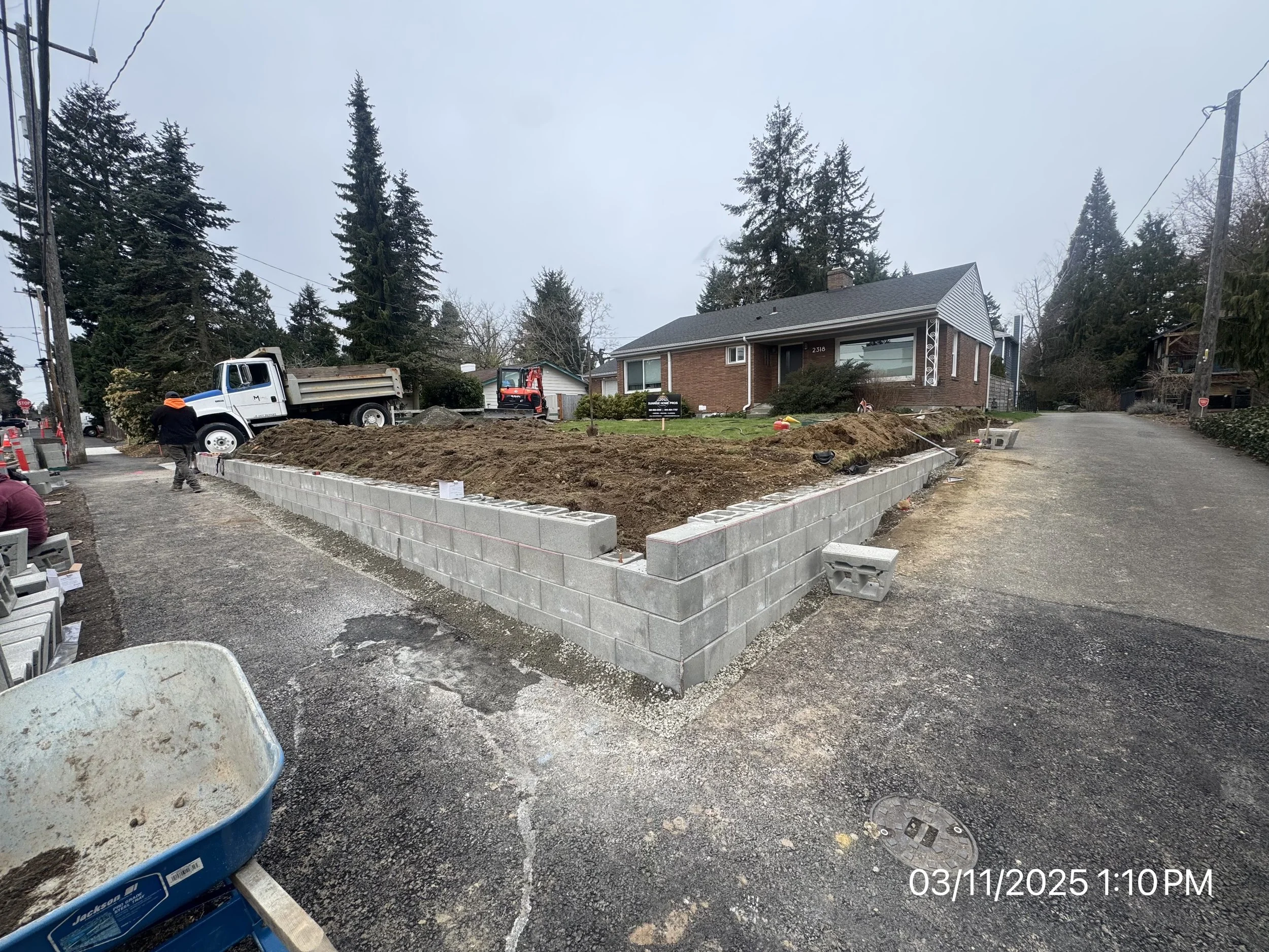 Construction site with a concrete block wall being built around a residential yard, with construction workers and equipment present.