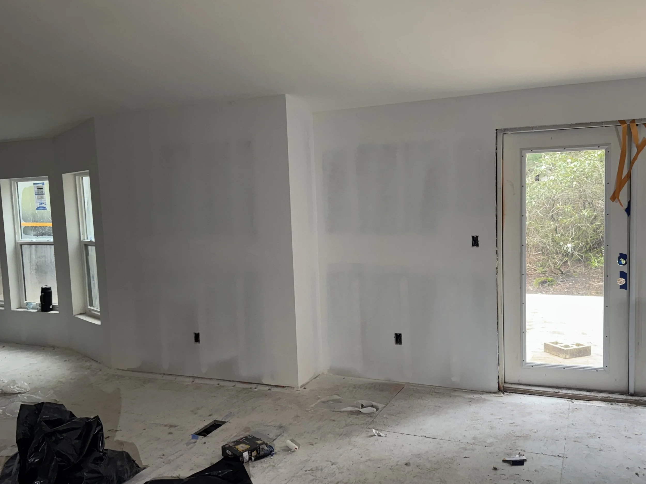 Interior of a room under construction with unfinished white walls, a door with a window, and construction tools and debris on the floor.