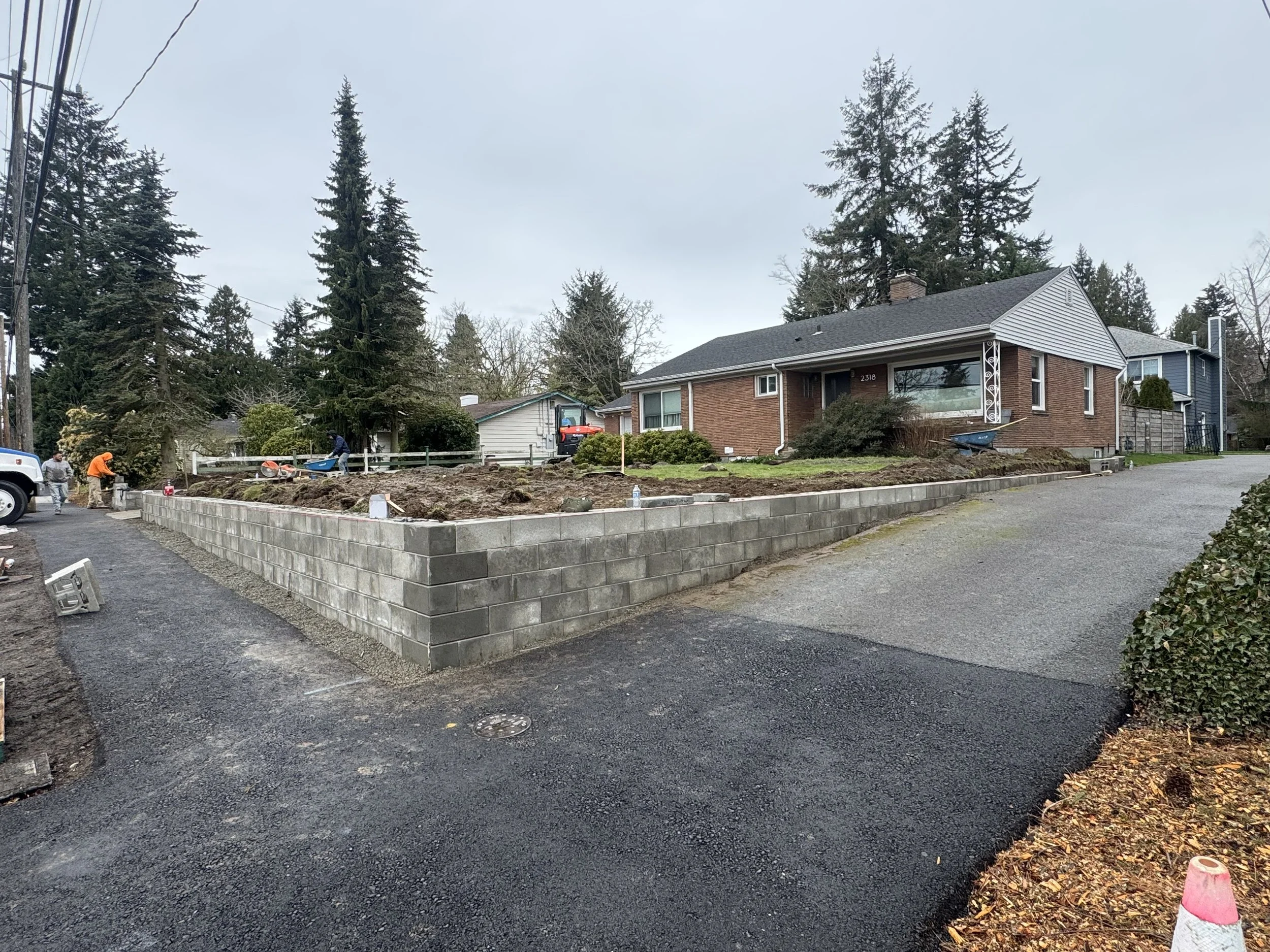 Construction workers building a brick retaining wall in front of a residential house on a cloudy day.
