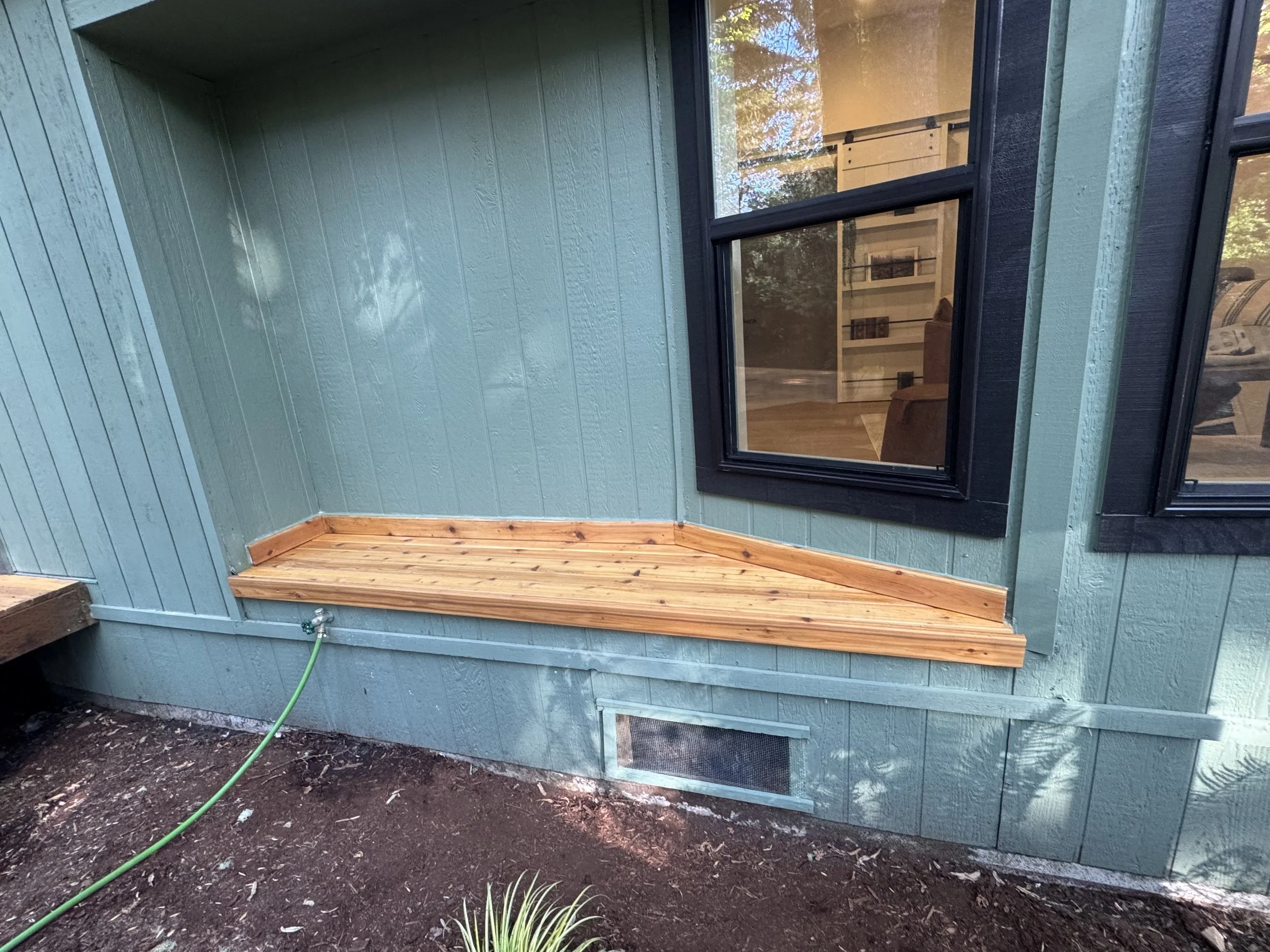 A backyard area with a wooden platform built against a green painted house wall, below a double window with black trim. A garden hose is connected near the bottom corner of the house, and the ground is bare soil. The inside of the house is visible th
