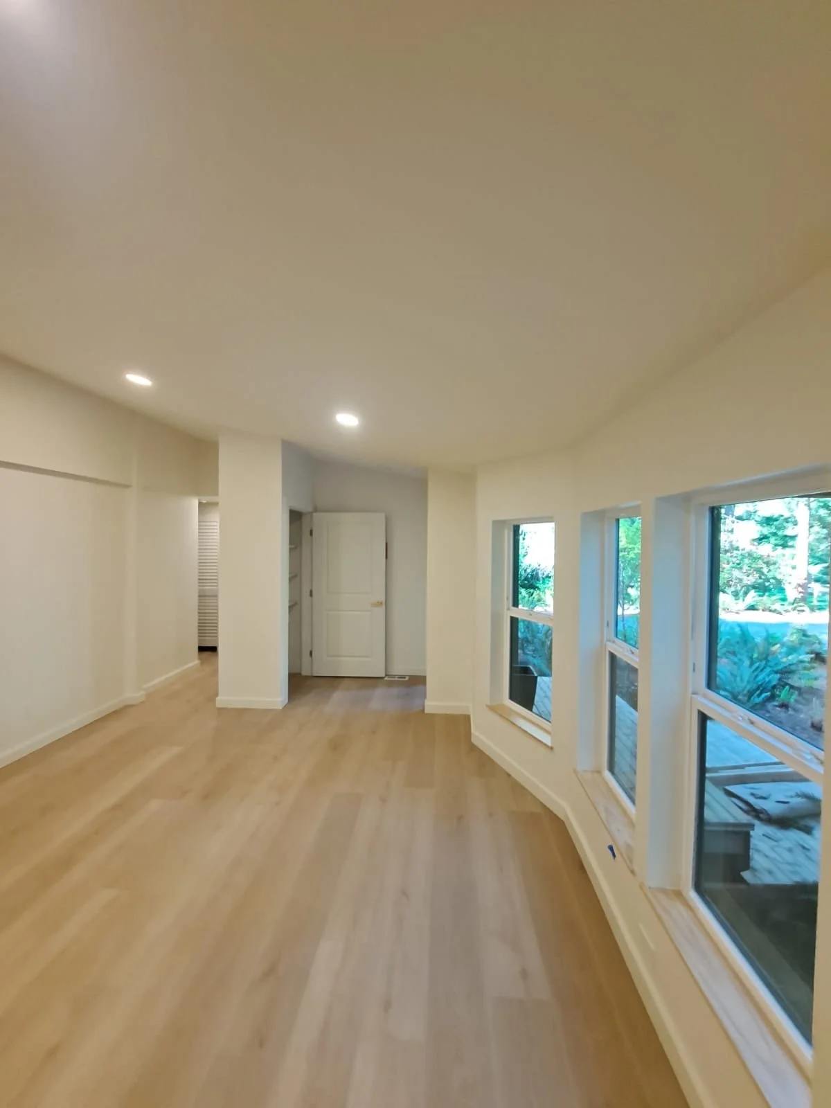 Empty room with large windows, light-colored hardwood flooring, white walls, and ceiling lights.