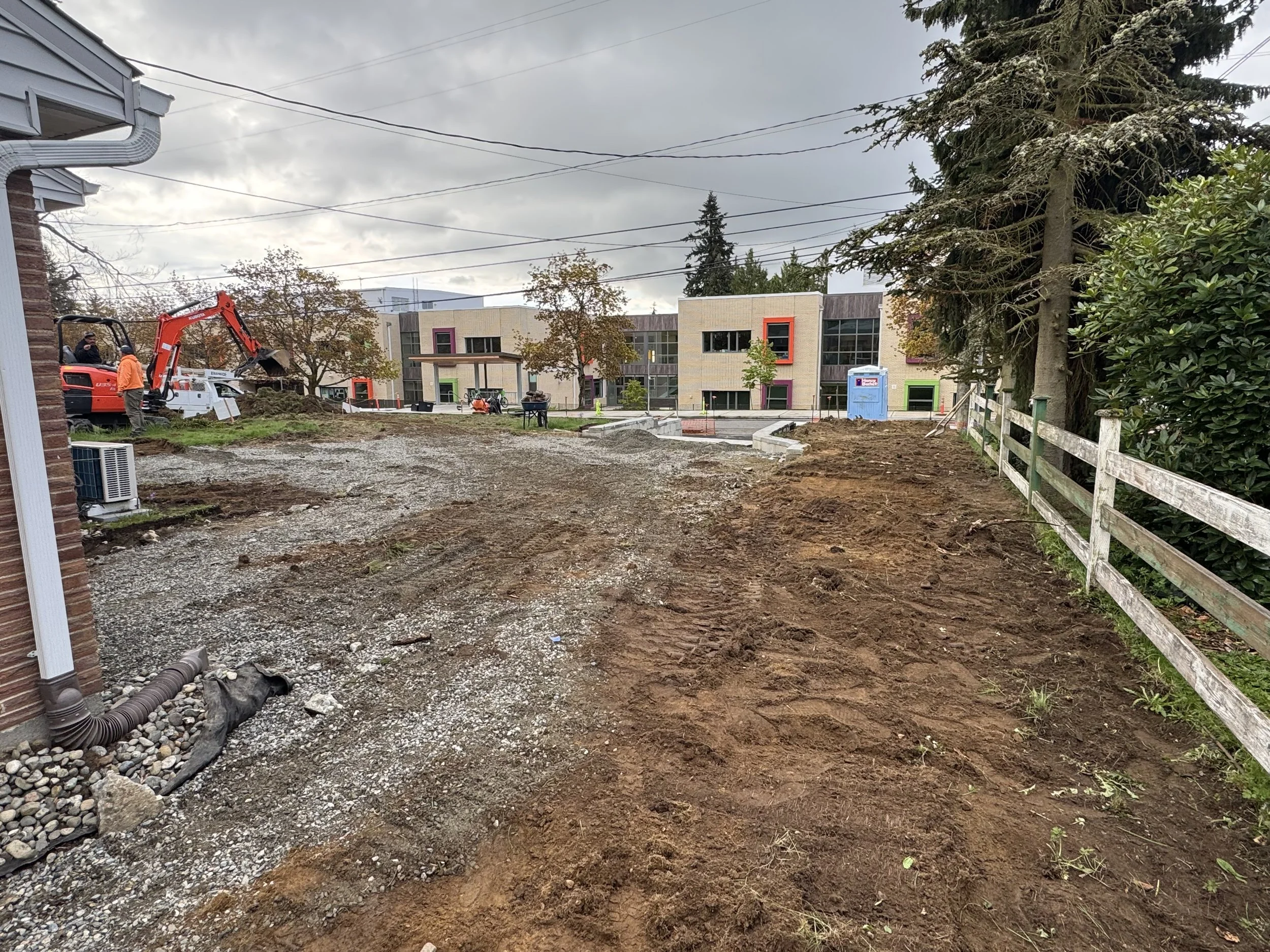 Construction site with dirt and gravel, a small excavator, workers, a portable toilet, and newly paved areas in front of modern buildings with colorful window frames.