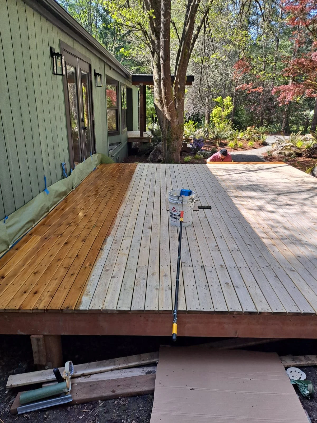 A wooden deck under construction next to a house with green siding, with a bucket, a measuring tape, and a tool on the deck, surrounded by trees and plants.