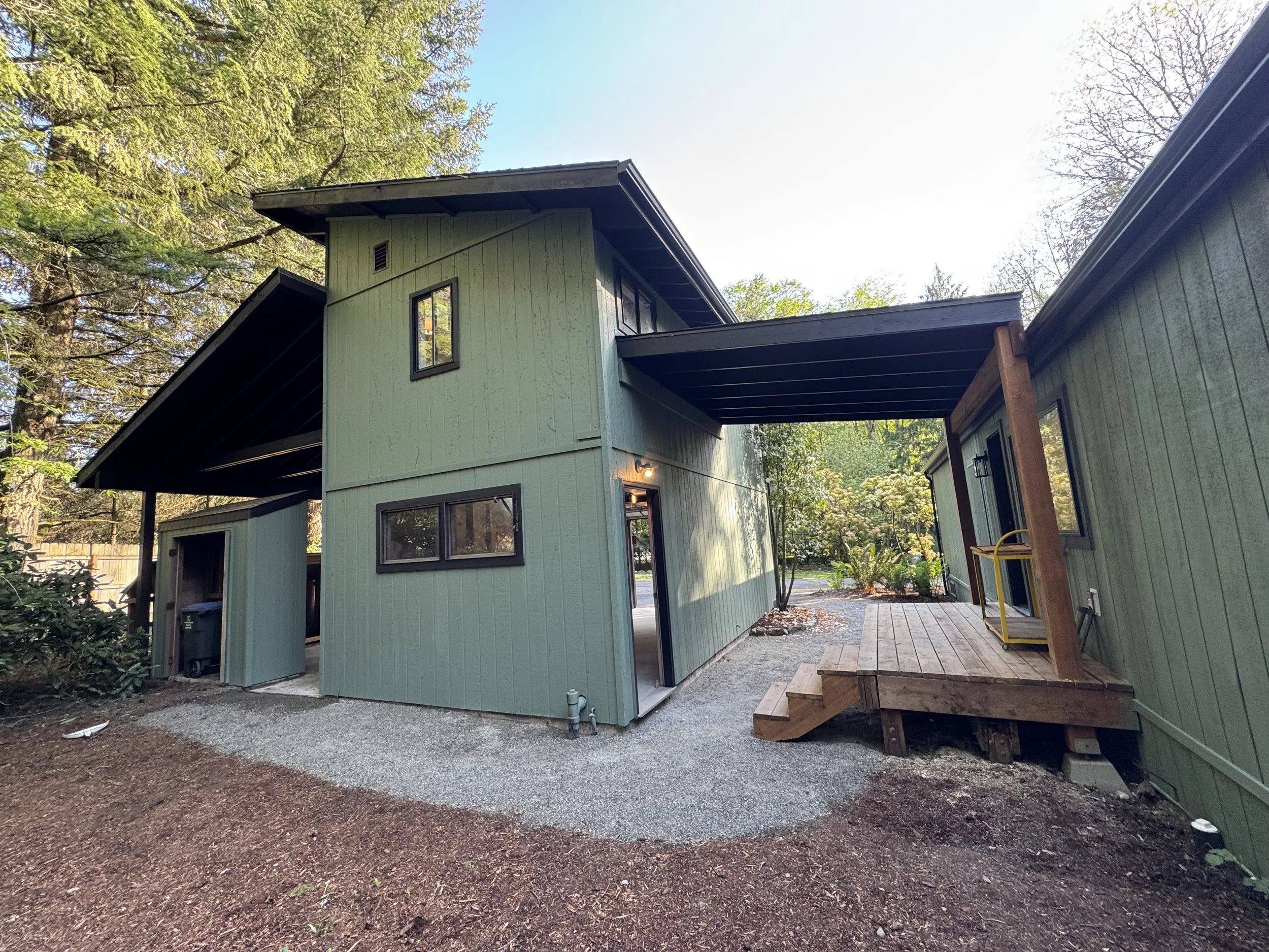 A green modern house with black-framed windows, a small wooden porch with stairs, surrounded by trees and a gravel patio, under a clear sky.