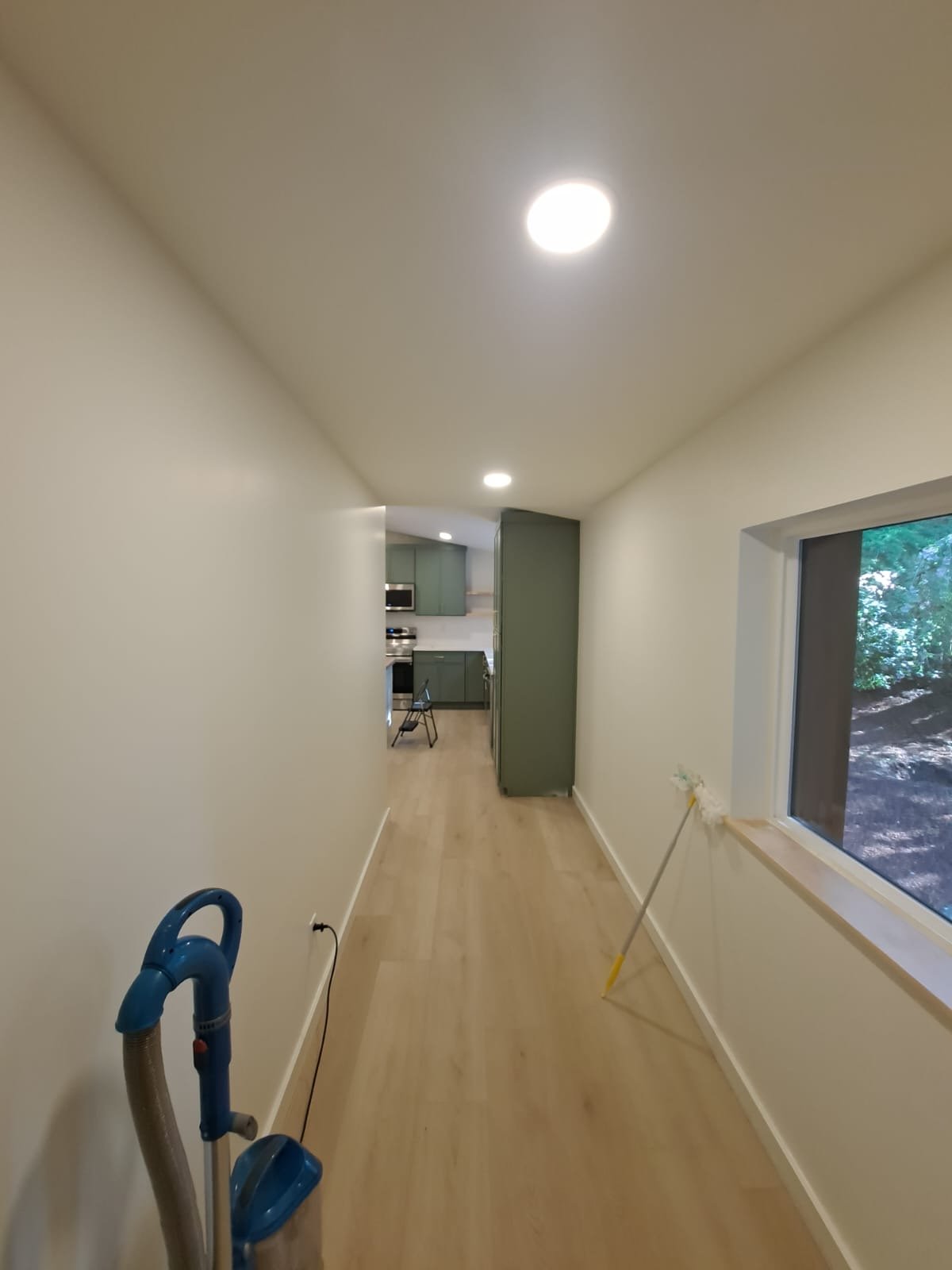 Empty hallway with light wood flooring, white walls, and ceiling lights, leading to a kitchen area with green cabinets and a window showing an outdoor scene.