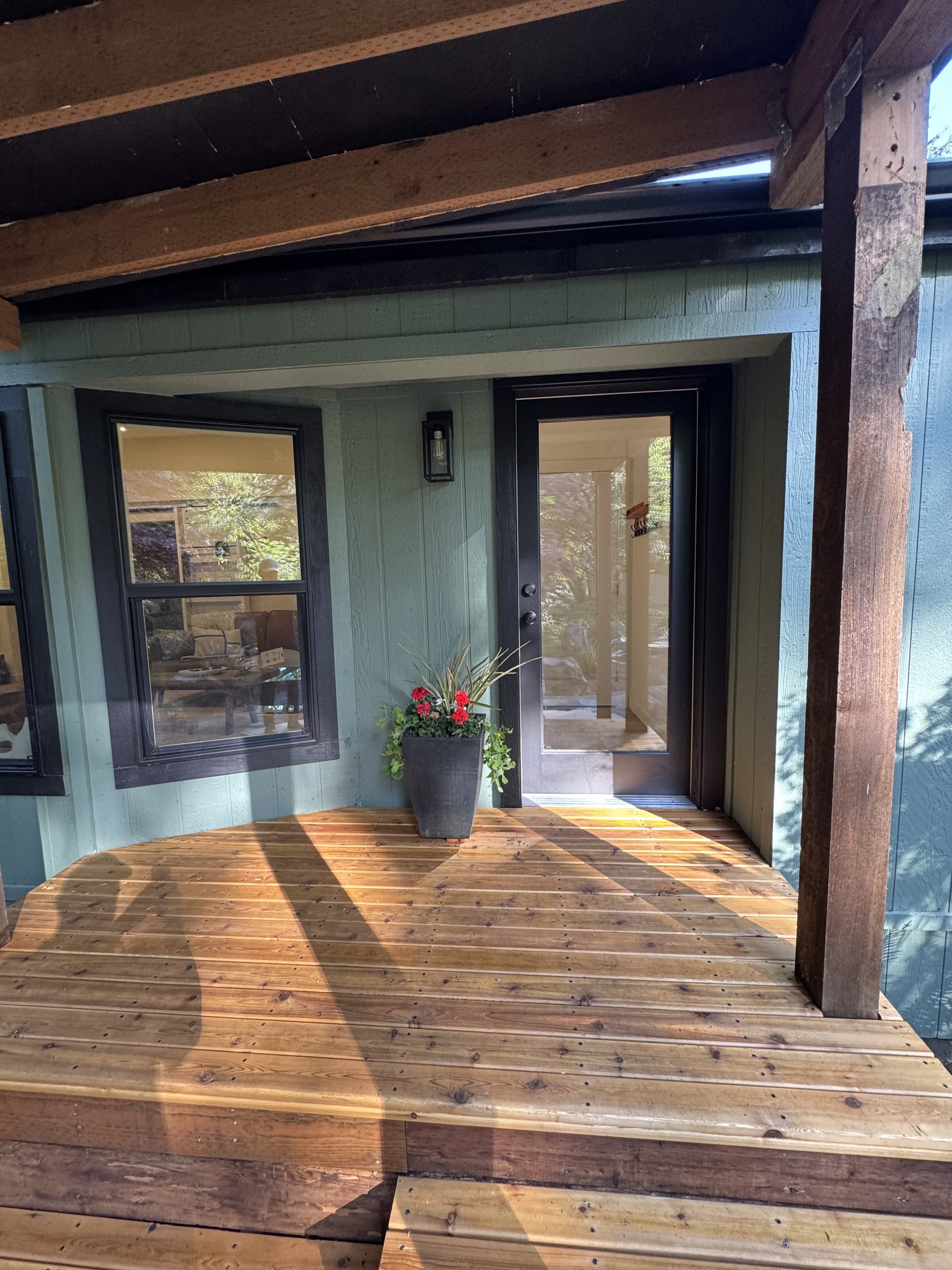 A close-up of a wooden porch with a potted plant with red flowers and green foliage, a black door with a glass panel, and two windows with black frames, attached to a building with green siding.