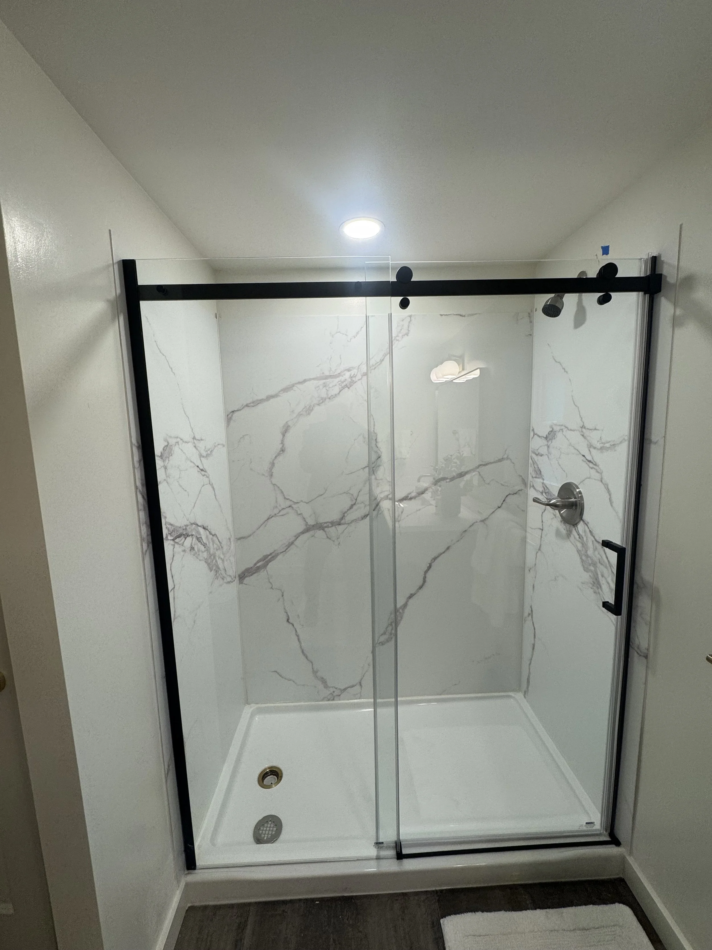 Empty modern shower with marble walls, glass door, and white floor, located in a bathroom with white walls and a wooden floor mat.