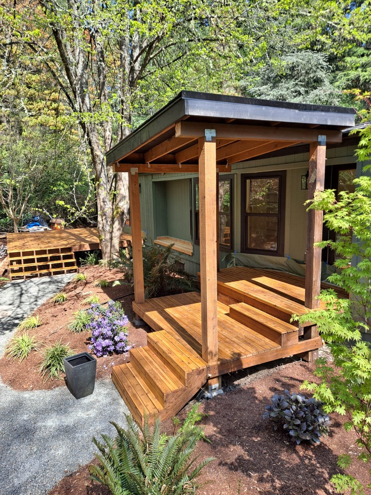 Wooden porch under construction in a garden with trees and plants, completed with steps, beams, and a roof, attached to a house with green siding and windows.