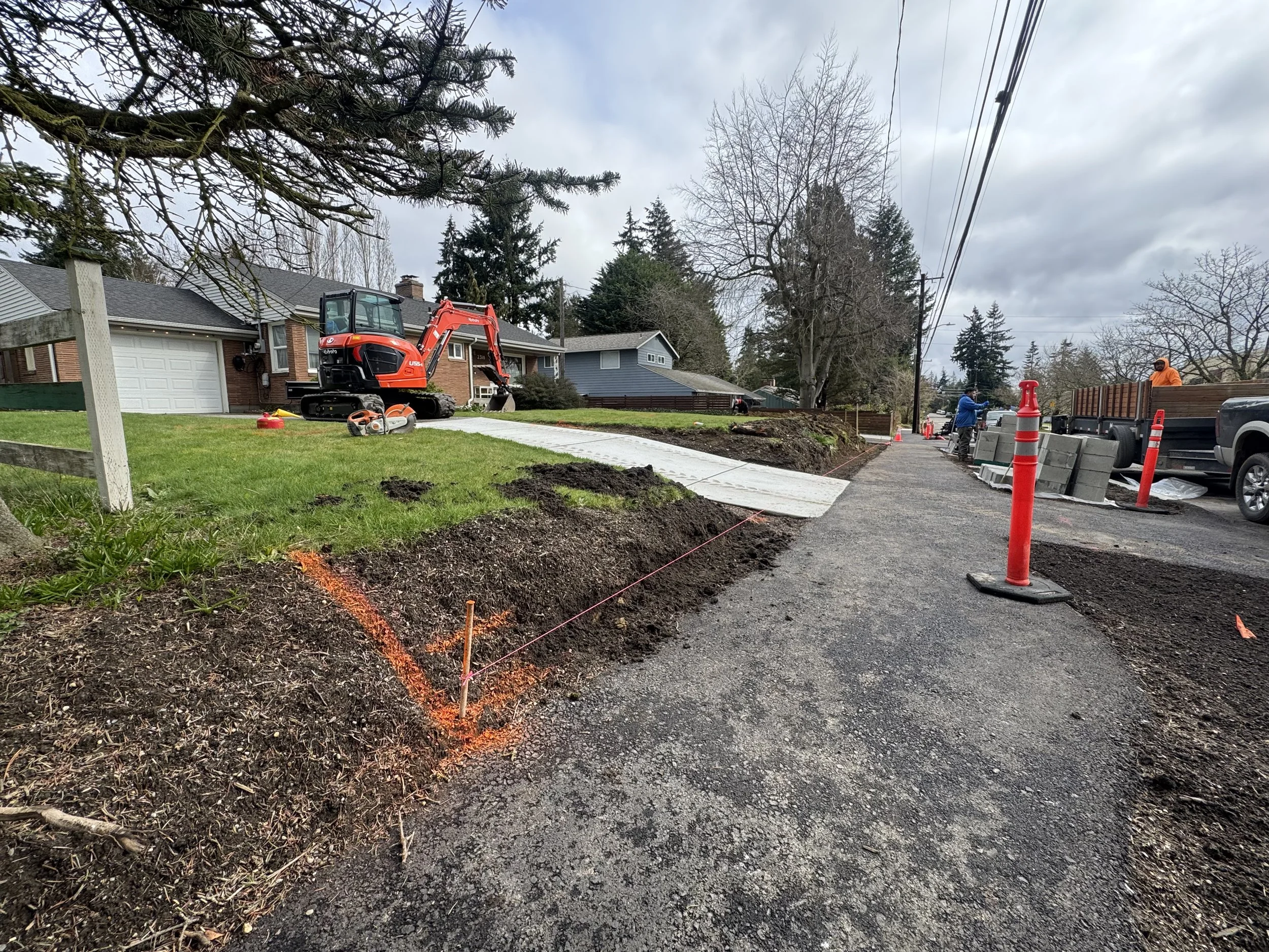 Construction workers installing a sidewalk in a residential neighborhood. A small orange excavator is on the lawn. Orange cones and barriers mark the work area.