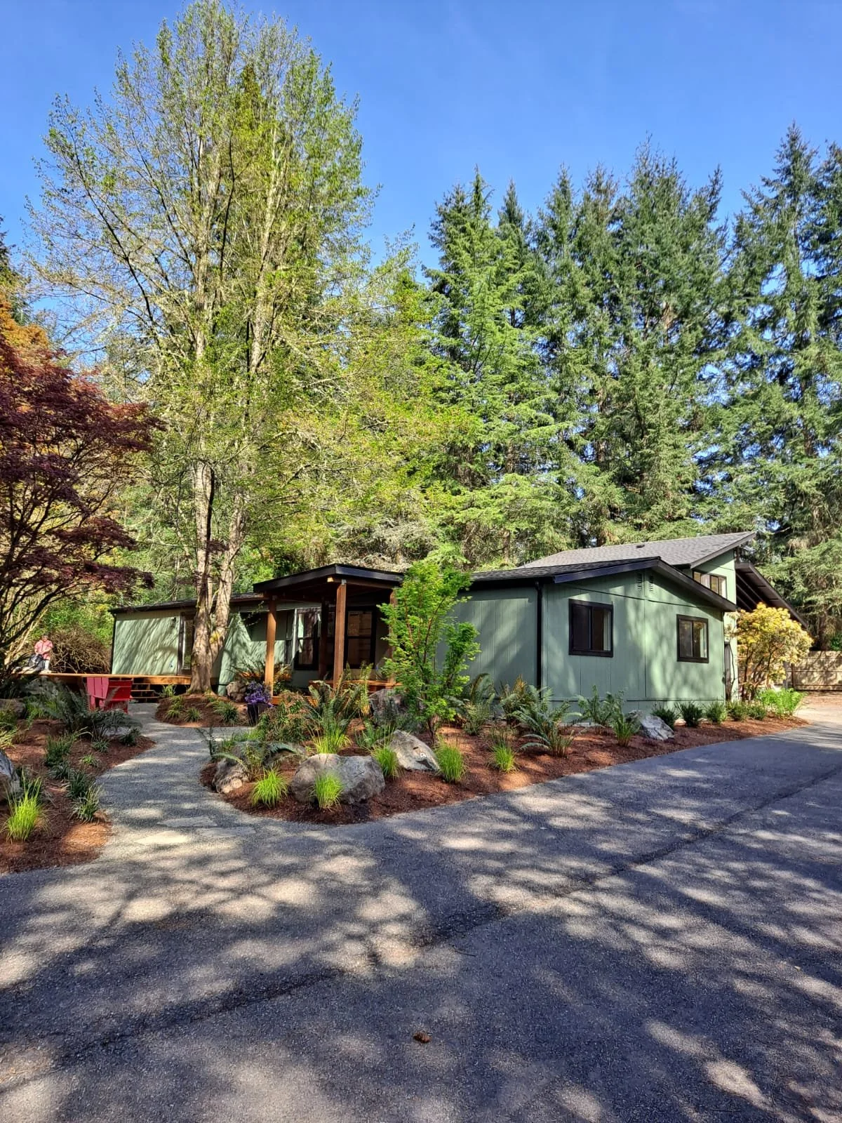 A light green house with black window frames surrounded by tall green trees, landscaped garden with rocks, shrubs, and a path, under a clear blue sky.