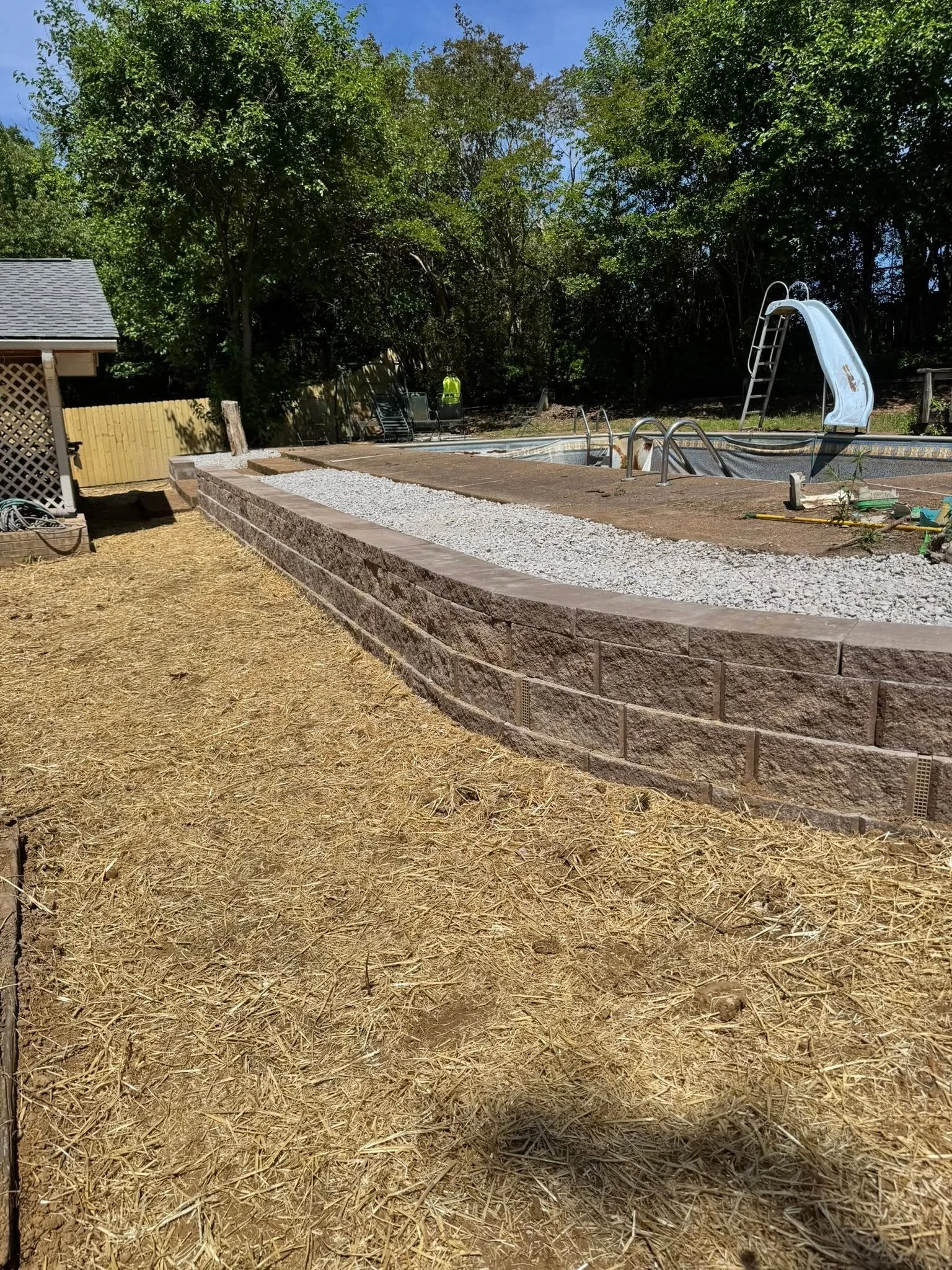 Backyard with a pool, slide, and surrounding trees, with some construction materials and tools nearby.