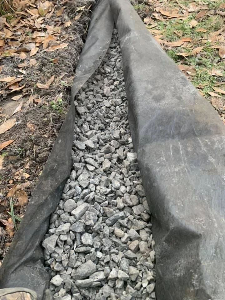 A trench lined with black fabric containing a layer of gravel on top, surrounded by fallen leaves and grass.