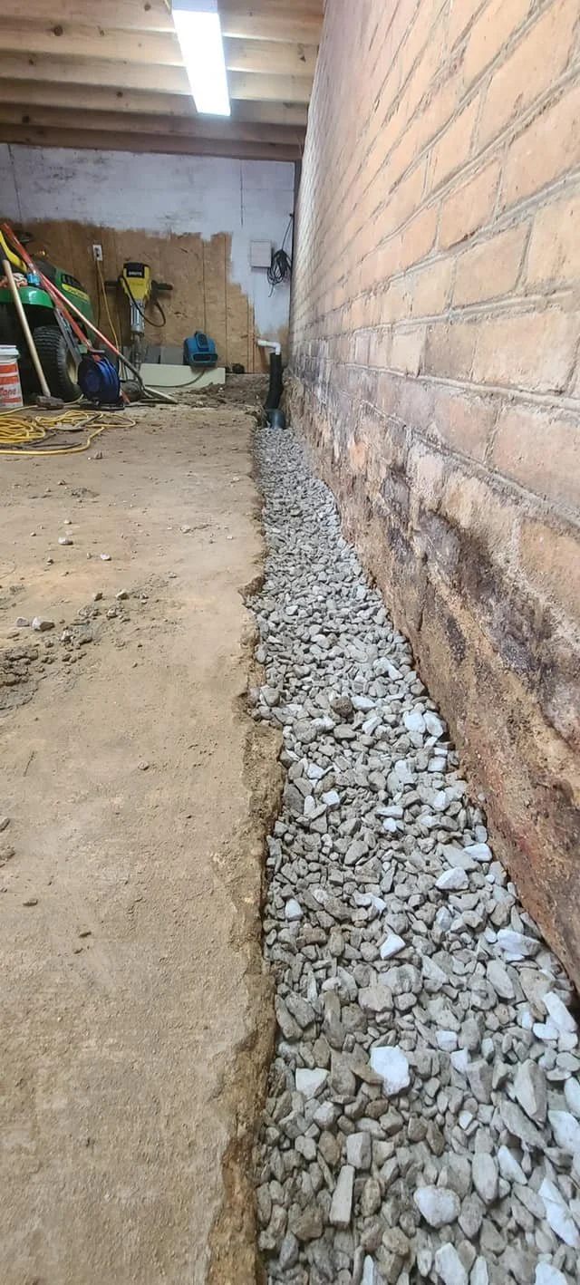A construction area with a trench filled with gravel along a brick wall, with tools and equipment visible in the background.