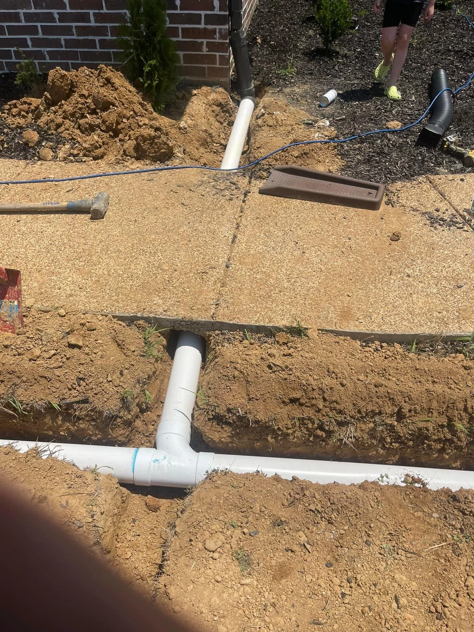 Under construction site showing PVC underground piping, a concrete sidewalk, tools, and a person in the background wearing yellow shoes.