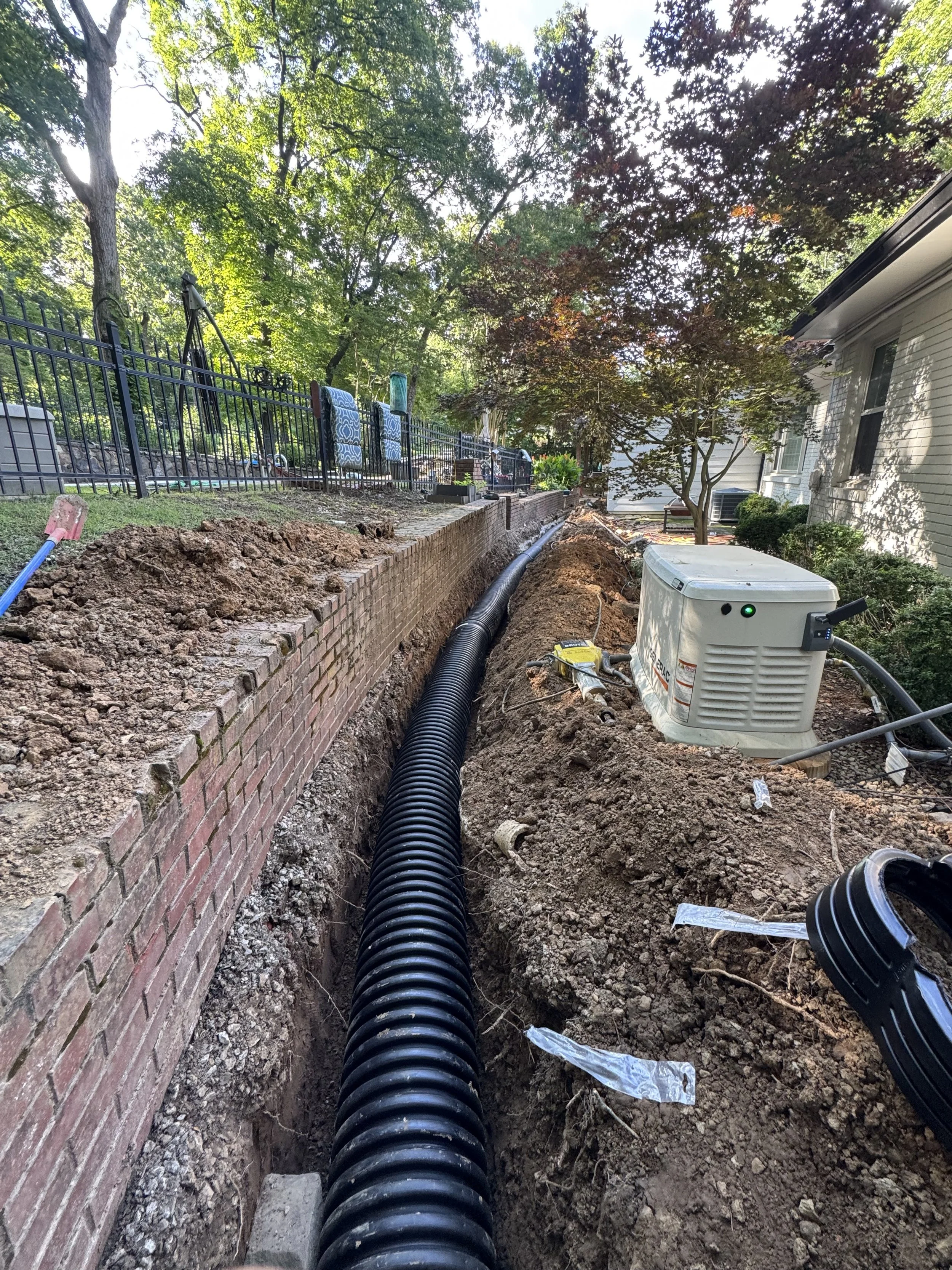 Construction site with a black corrugated pipe running along a trench, a white generator, and a brick wall on the left, in a residential backyard with trees and a house on the right.