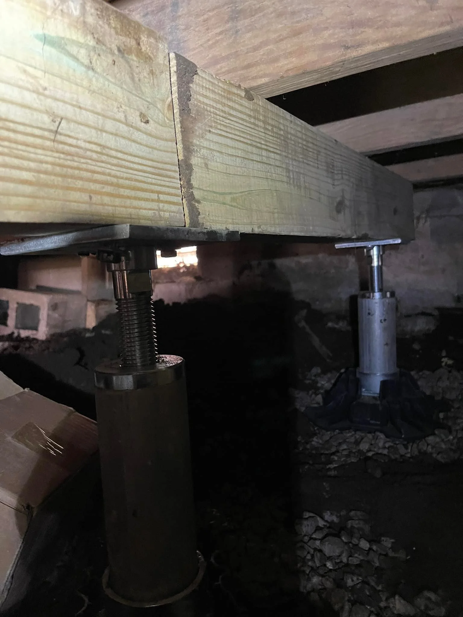 Underneath a wooden floor, metal adjustable support jacks are holding the floor in place. The support jacks sit on rough gravel ground.