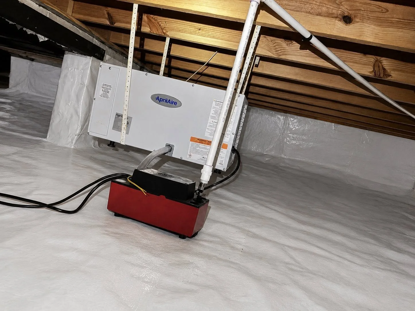 An Aprilaire attic air purifier installed in an unfinished attic space with wooden ceiling beams and insulation on the floor.