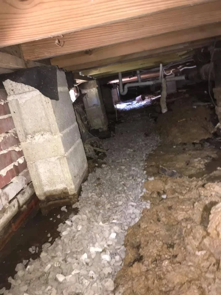 Underground crawl space with exposed dirt, gravel, concrete block foundation, and plumbing pipes running along the ceiling.