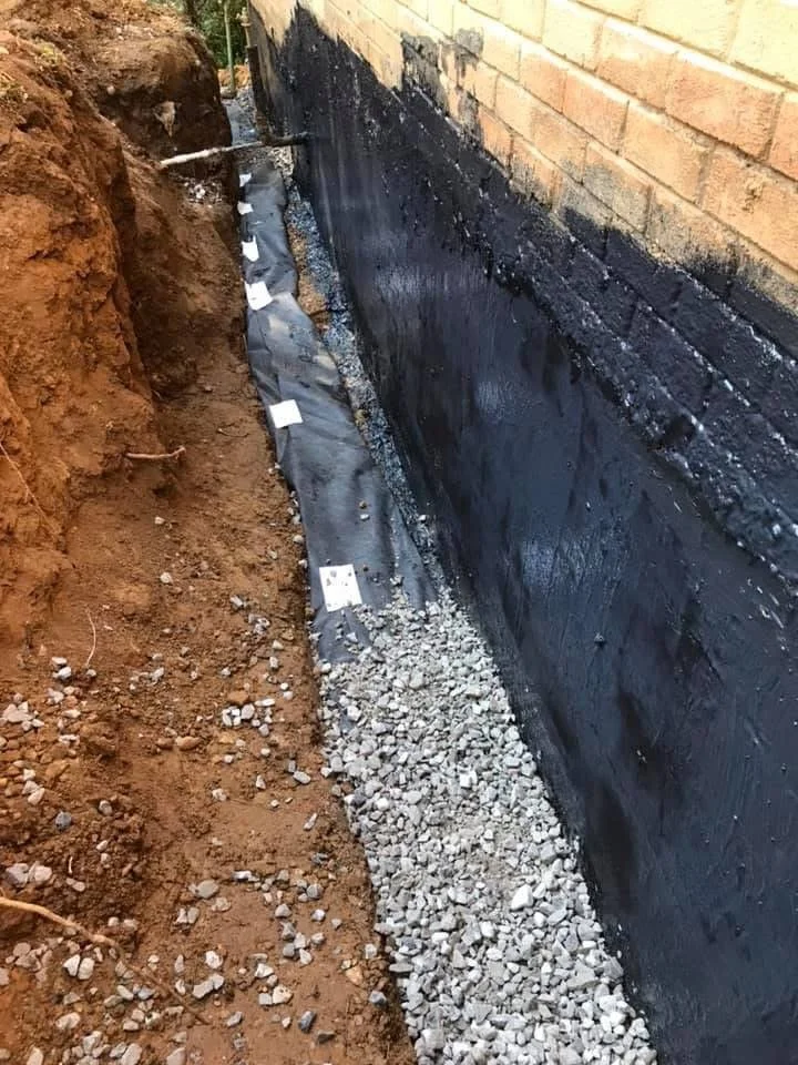 Excavated trench along the side of a brick wall with black waterproofing membrane, drainage gravel, and a pipe, indicating foundation waterproofing work.