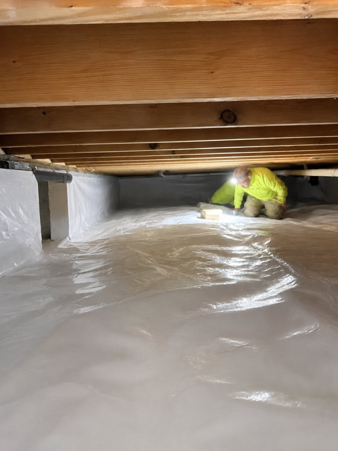 Worker in a yellow jacket installing a vapor barrier on the floor of a crawl space with wooden beams overhead.