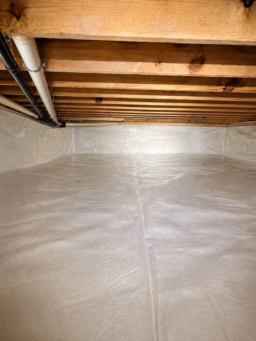A basement corner with wooden ceiling beams, black and white pipes, and a white vapor barrier on the walls and floor.