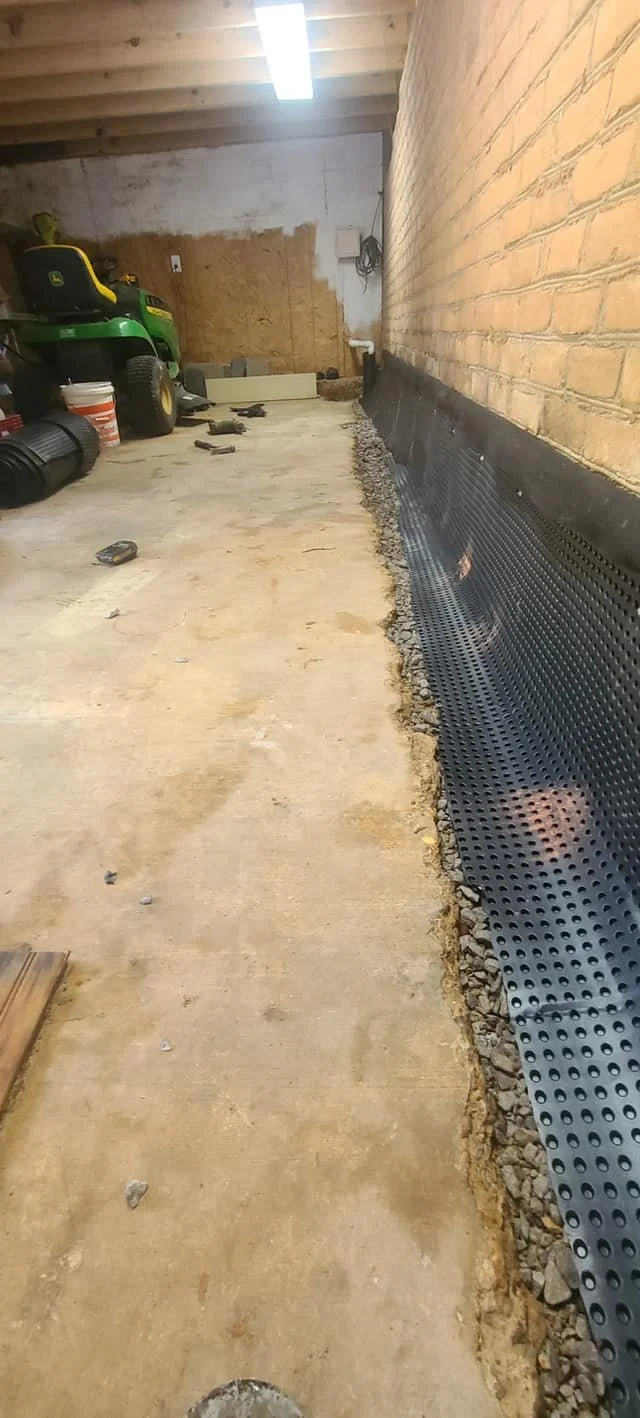 Construction in progress on a concrete floor next to a brick wall with black drainage membrane installed along the base.