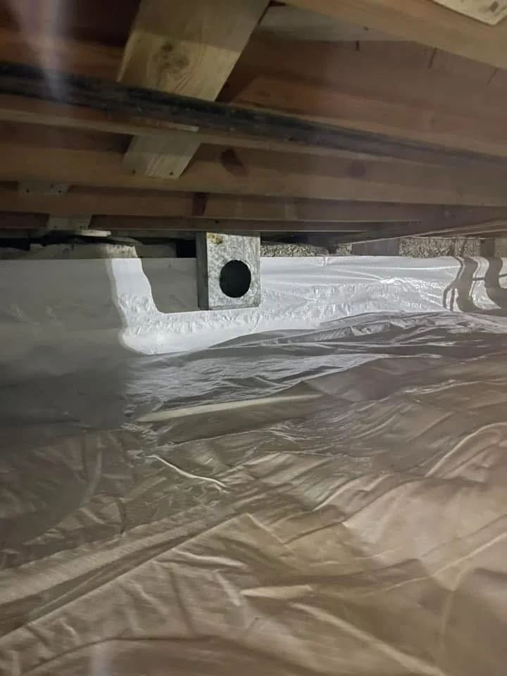 The image shows the underside of a wooden structure, possibly a deck or porch, with a metal bracket fixing the wood to a concrete or wooden surface. There is a white vapor barrier or plastic sheeting covering the ground underneath.