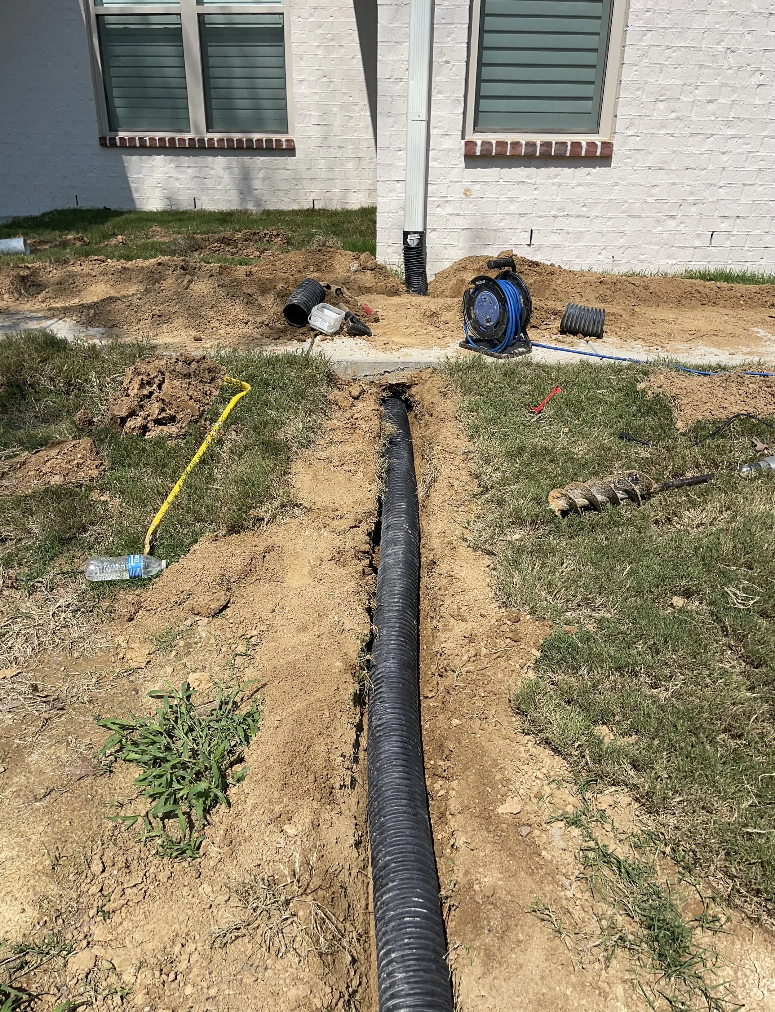 A trench with a black corrugated pipe installed runs from the foreground to the background. Construction tools, a water bottle, and outdoor equipment are scattered around a grassy area in front of a house with white brick walls and green window shutters.