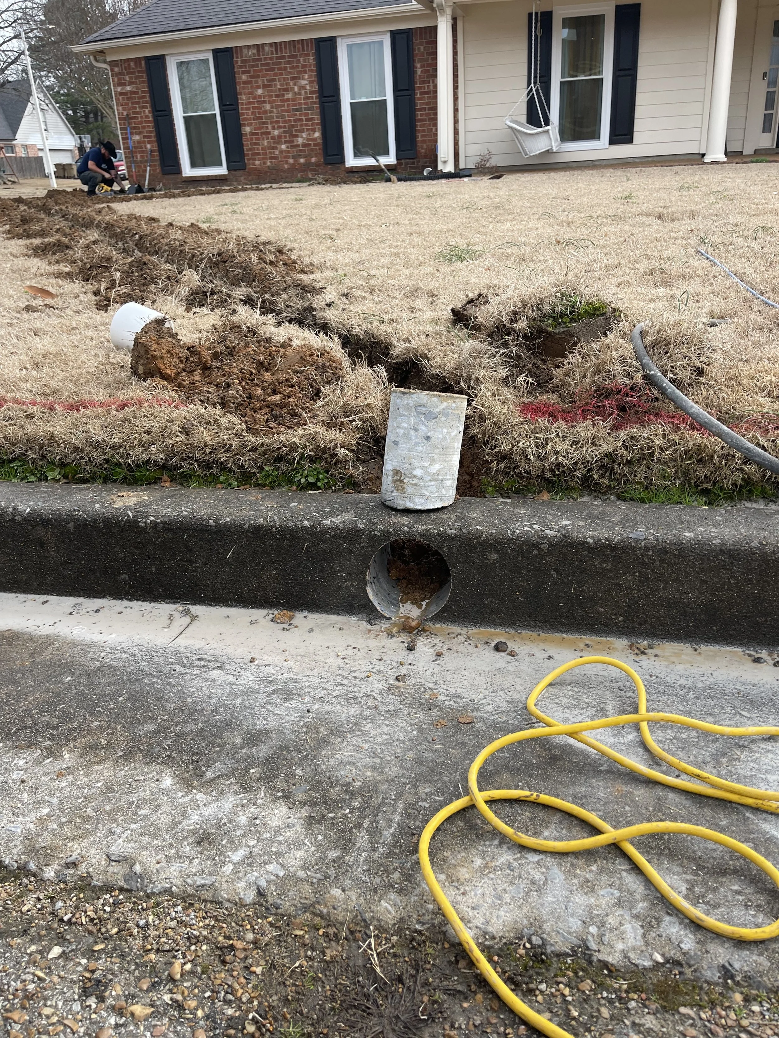 A lawn renovation process showing a trench dug along the edge of the driveway, with dirt and roots piled up, a pipe opening in the curb, and construction tools including a yellow extension cord and a white cylindrical object. A house with brick and siding, a swing, and a person working are visible in the background.