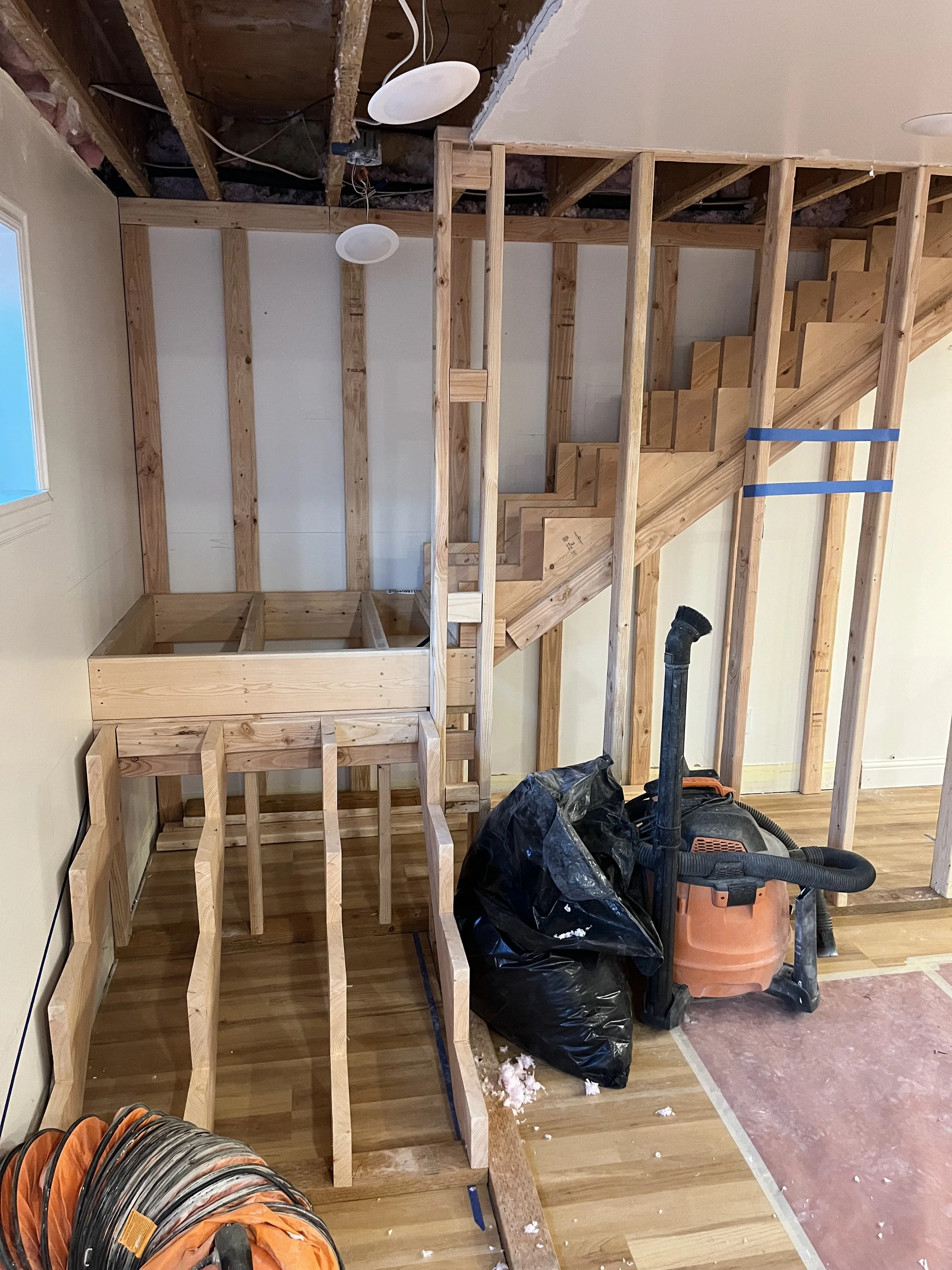 Interior of a house under renovation showing exposed wooden staircase framing, construction tools, and debris.