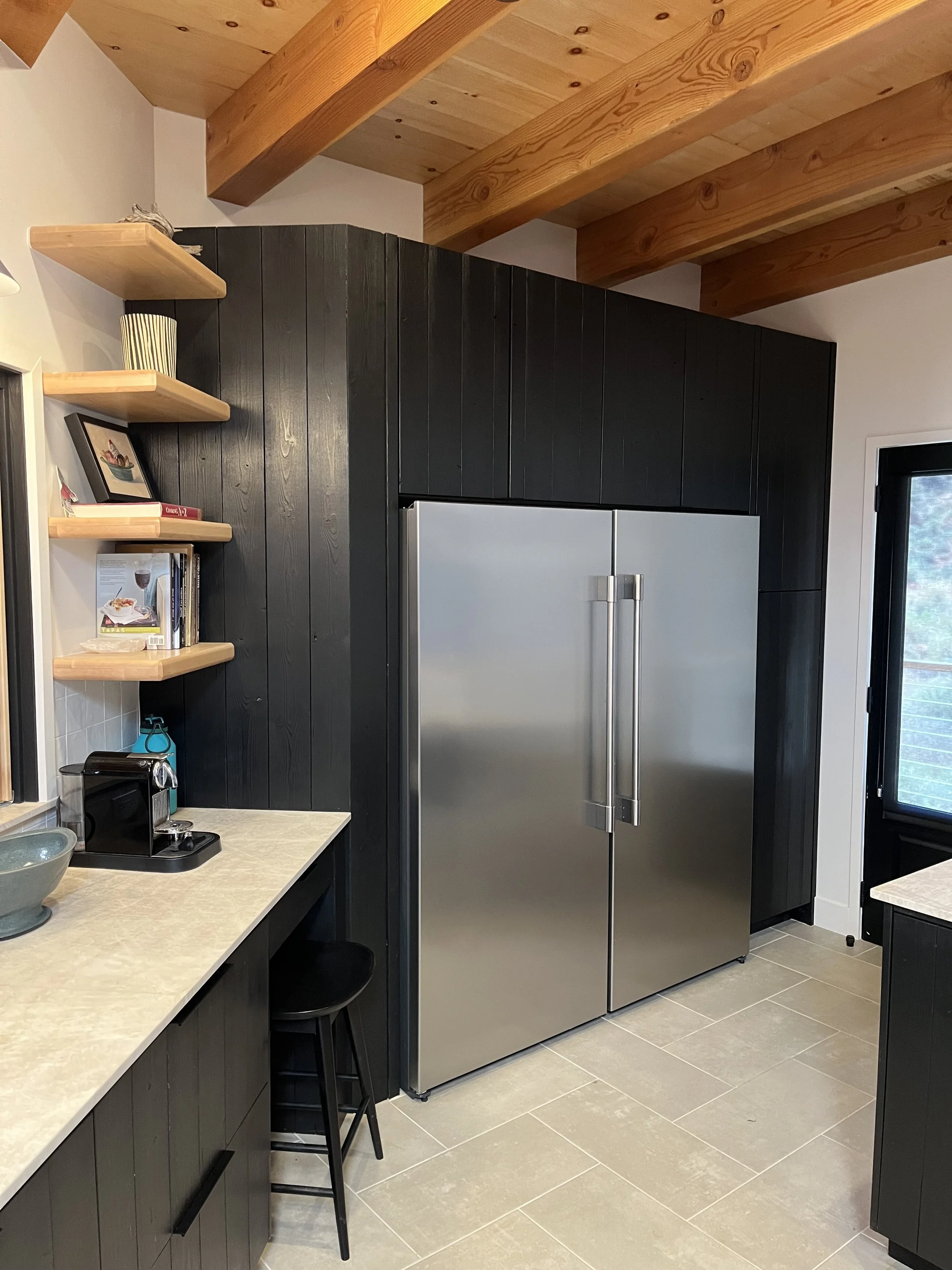 Modern kitchen with black cabinets, a silver refrigerator, wooden shelves, and a countertop with a coffee machine and a bowl.