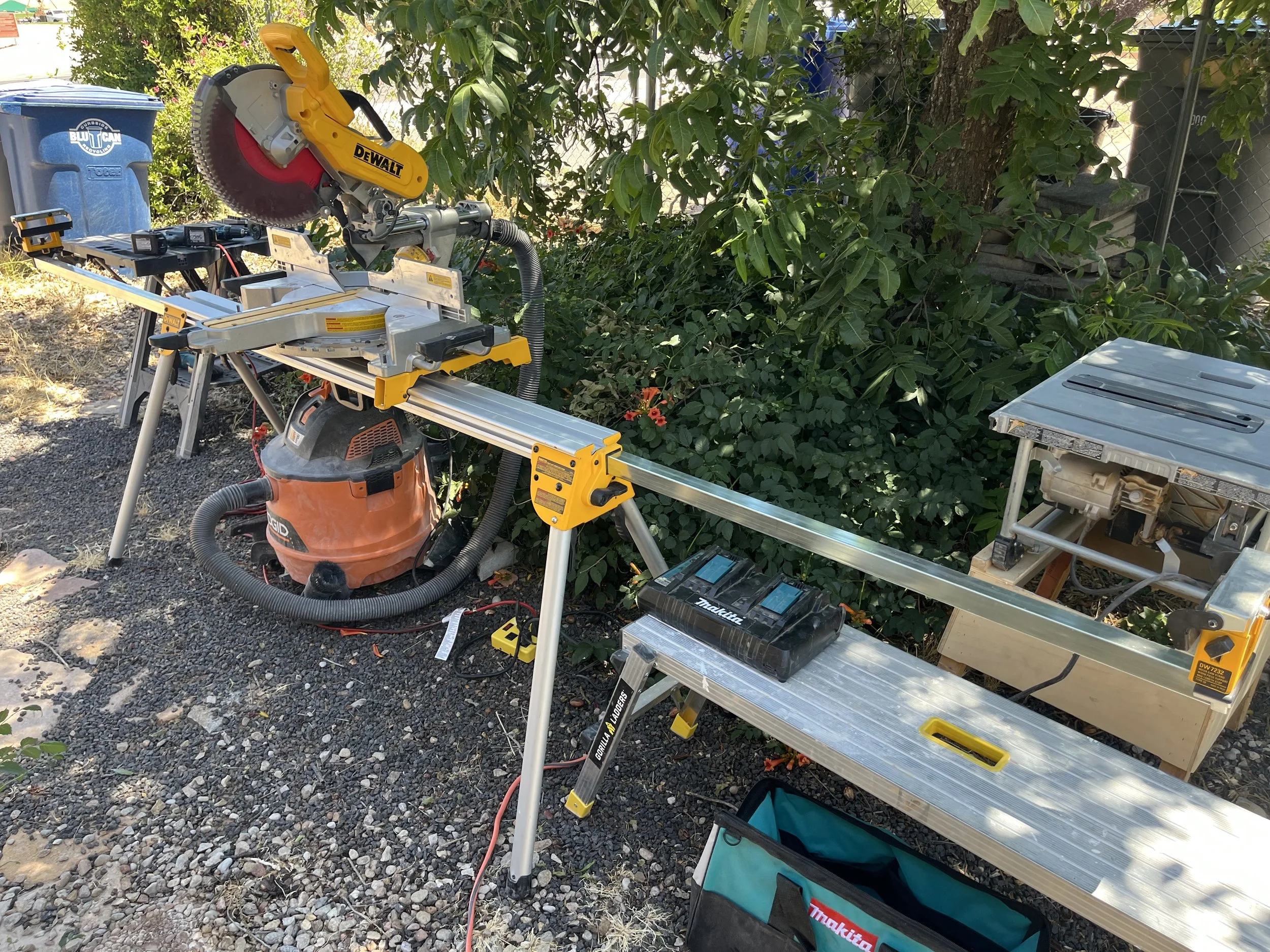 A workspace outdoors with power tools and equipment, including a large circular saw, work tables, a toolbox, and a trash bin, surrounded by green trees and plants.