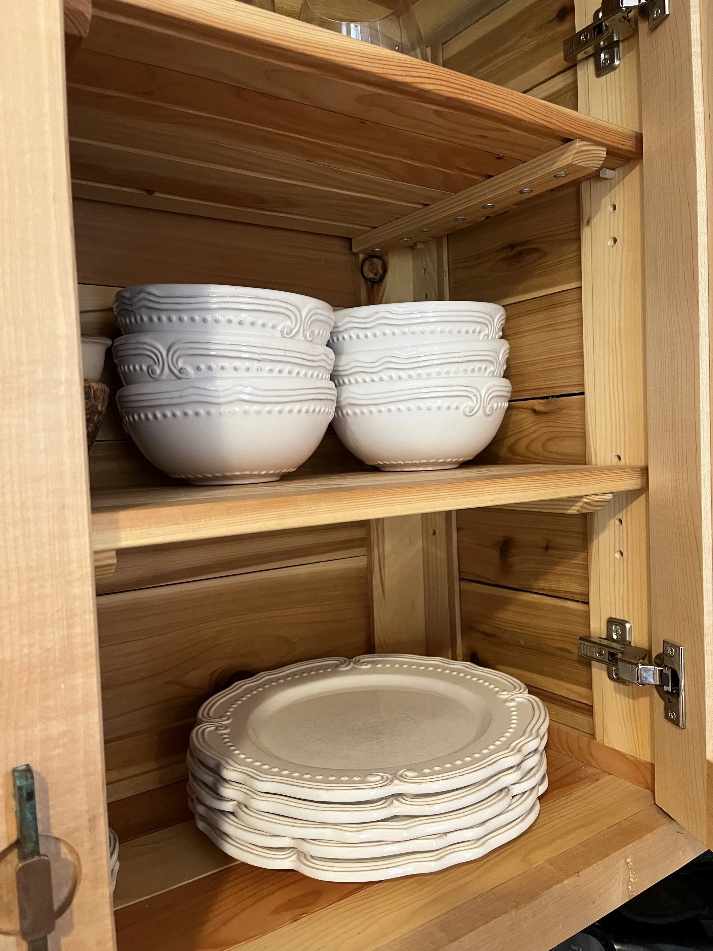 Open kitchen cabinet revealing stacked white ceramic plates and bowls inside a wooden cabinet with hinges.