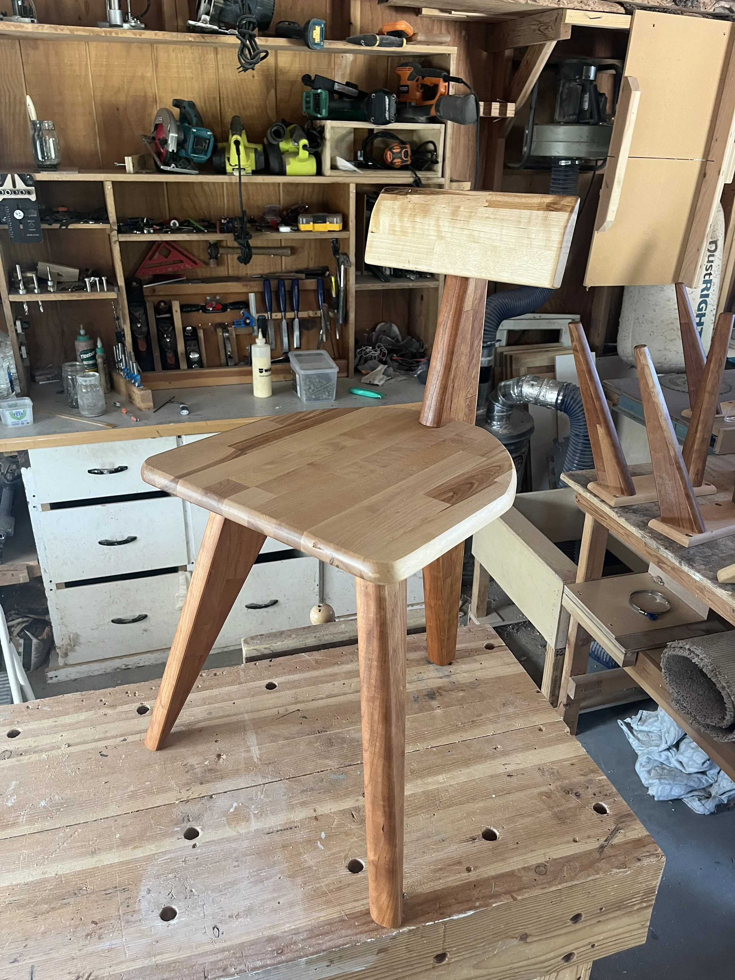 A woodworking workspace with a partially assembled chair in the center, surrounded by woodworking tools, clamps, and supplies on the shelves and workbenches.