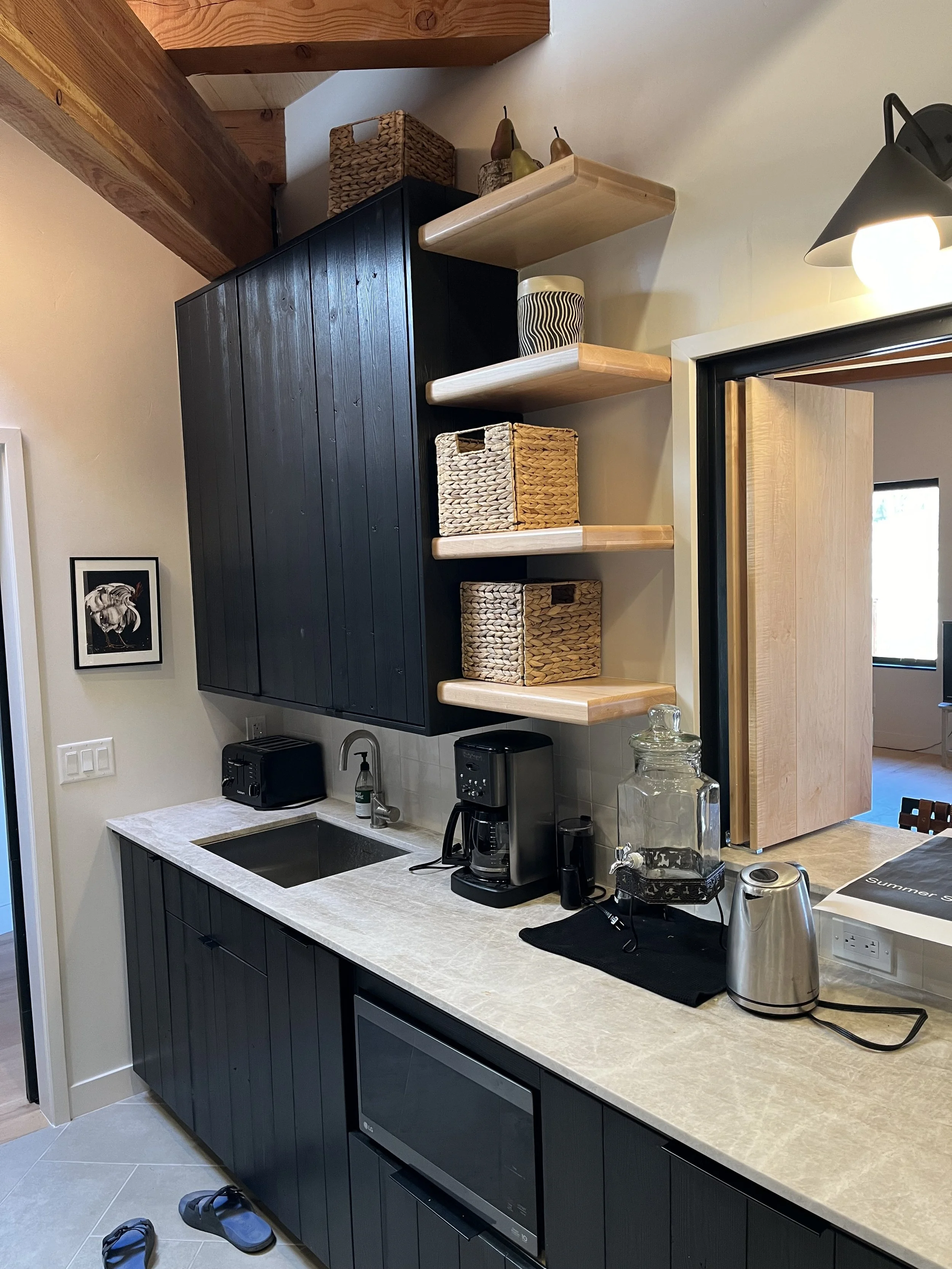 Kitchen with black cabinets, white countertop, coffee machine, water dispenser, and decor on open wooden shelves, with a doorway revealing another room.